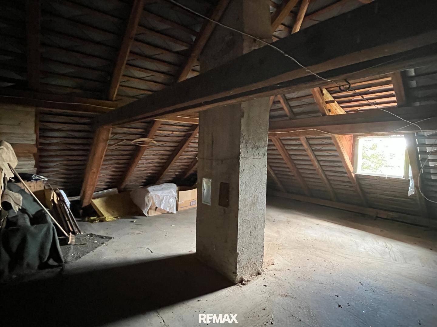 Geräumiger Dachboden mit Holzbalken, Betonboden und einem kleinen Fenster, das Licht spendet.