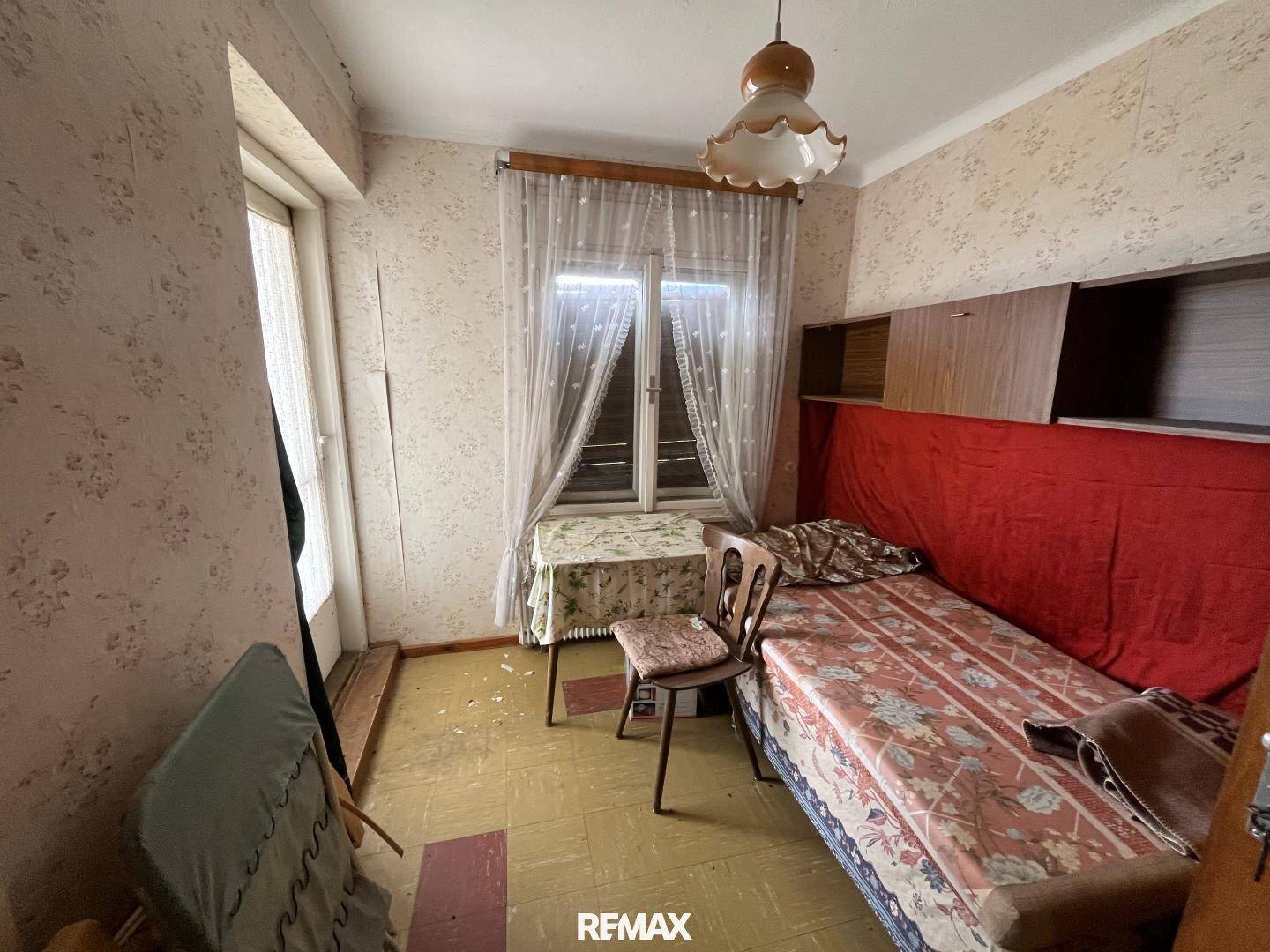 Altes Schlafzimmer mit gemusterter Tapete, Einzelbett und Fenster, das Tageslicht hereinlässt.