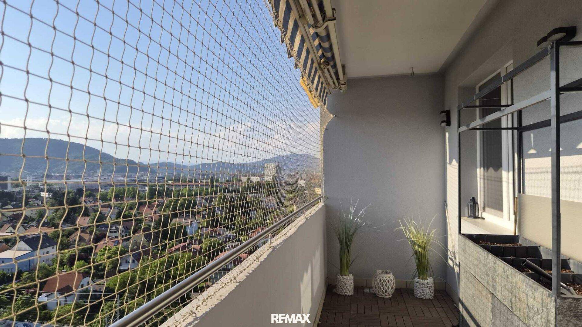 Sonniger Balkon mit Schutznetz, Holzboden und weitem Blick über die Stadt und die Berge.