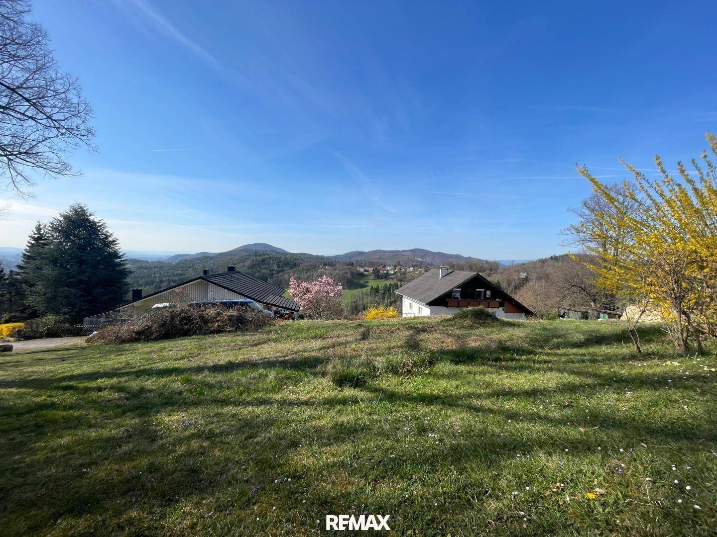 Panoramablick über eine sonnige Wiese mit mehreren Häusern und einer malerischen Bergkulisse.