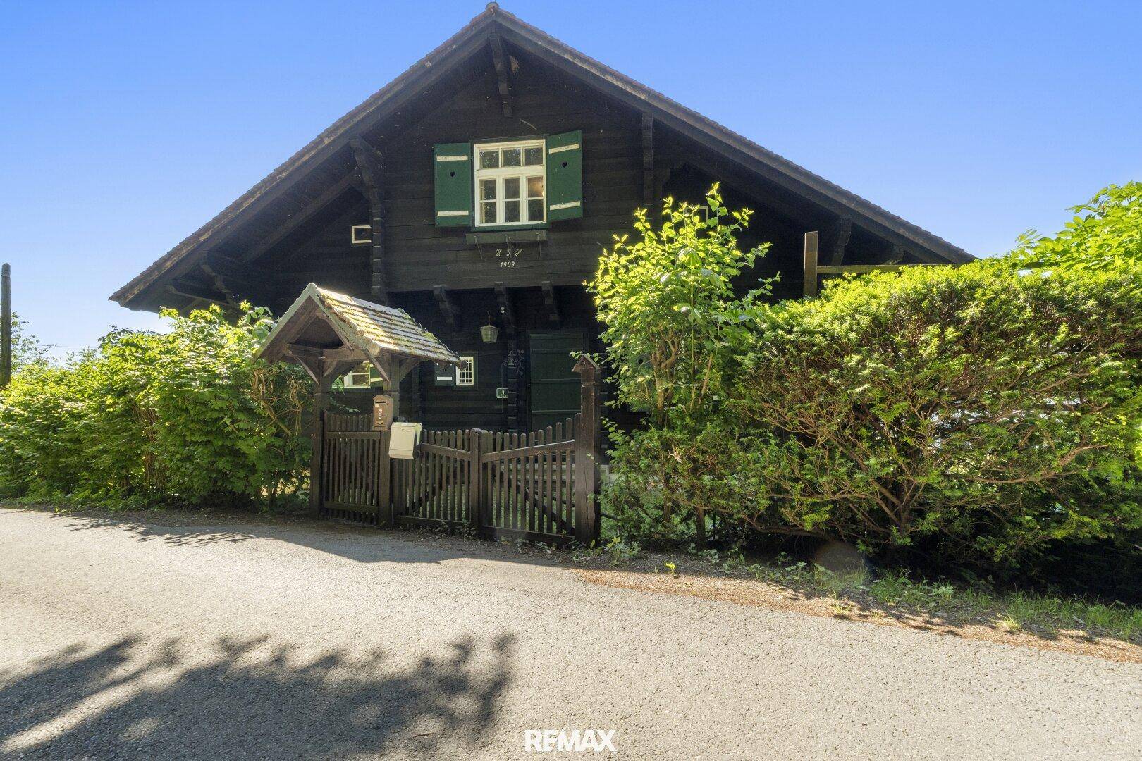 Traditionelles Holzhaus mit Holzzaun und grünen Fensterläden, umgeben von Hecken und Bäumen.