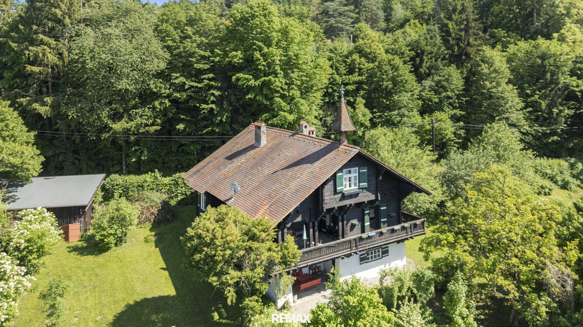 Historisches Holzhaus mit Satteldach und Balkon, eingebettet in eine grüne Hügellandschaft.