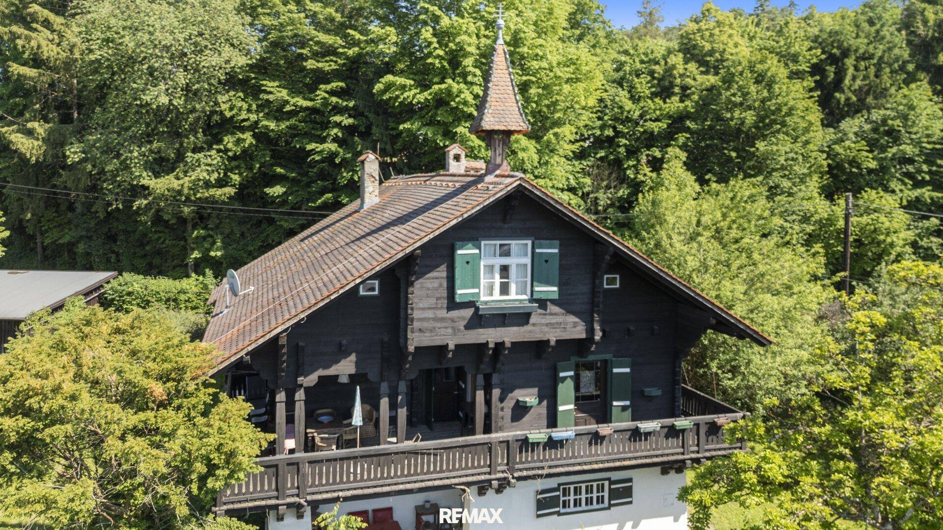 Charmantes Holzhaus mit traditionellem Balkon und grünen Fensterläden, umgeben von Bäumen.