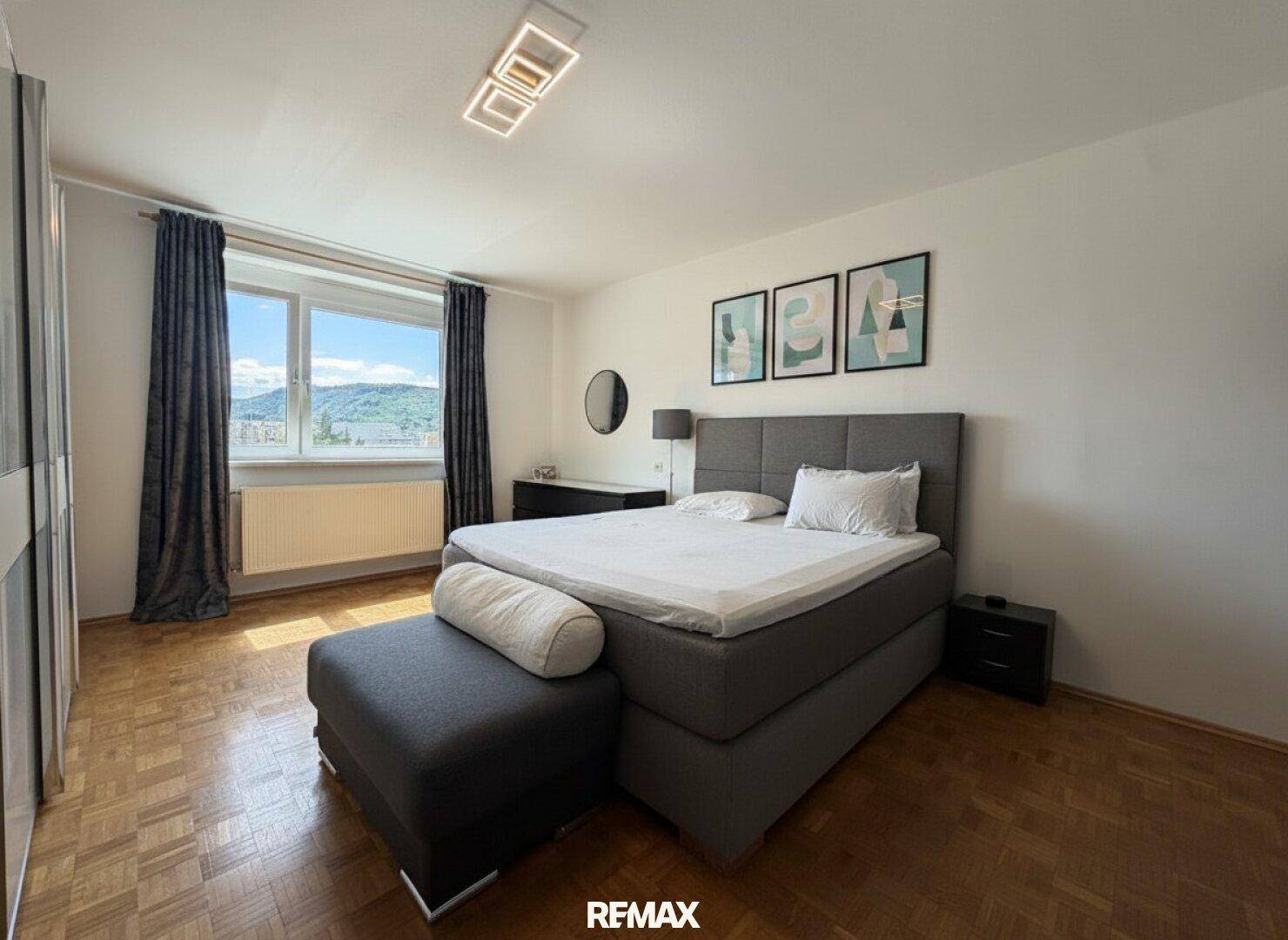 Schlafzimmer mit großem Bett, Parkettboden und Fenster mit Ausblick.