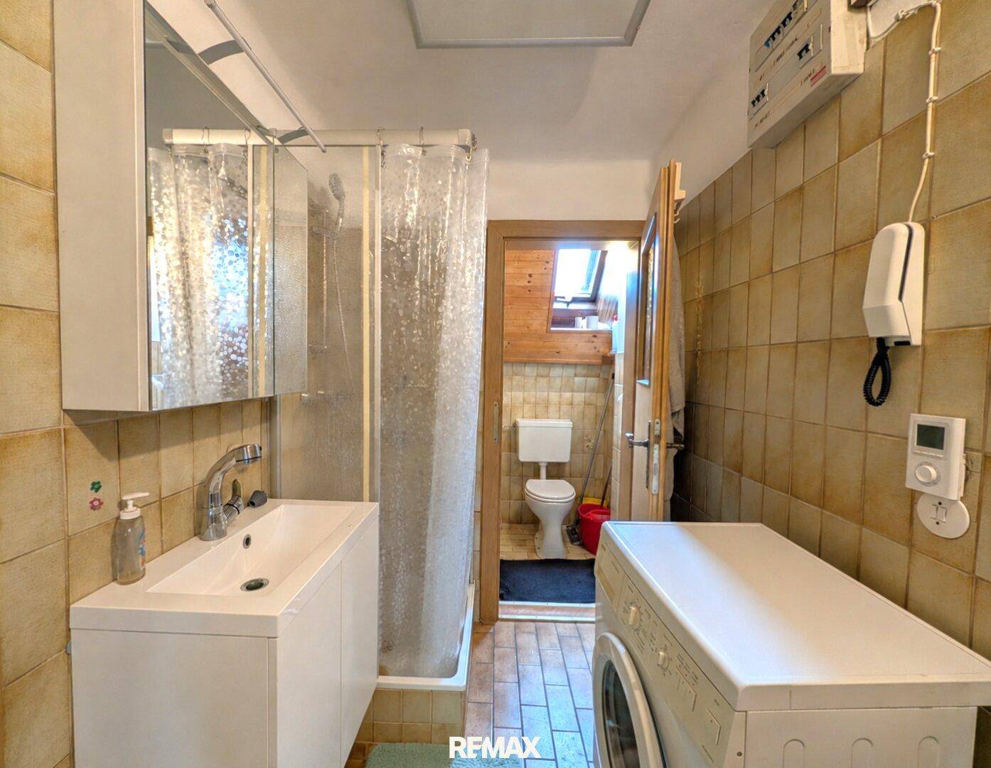 Badezimmer mit Dusche, Waschbecken und Waschmaschine, angrenzend an eine separate Toilette.