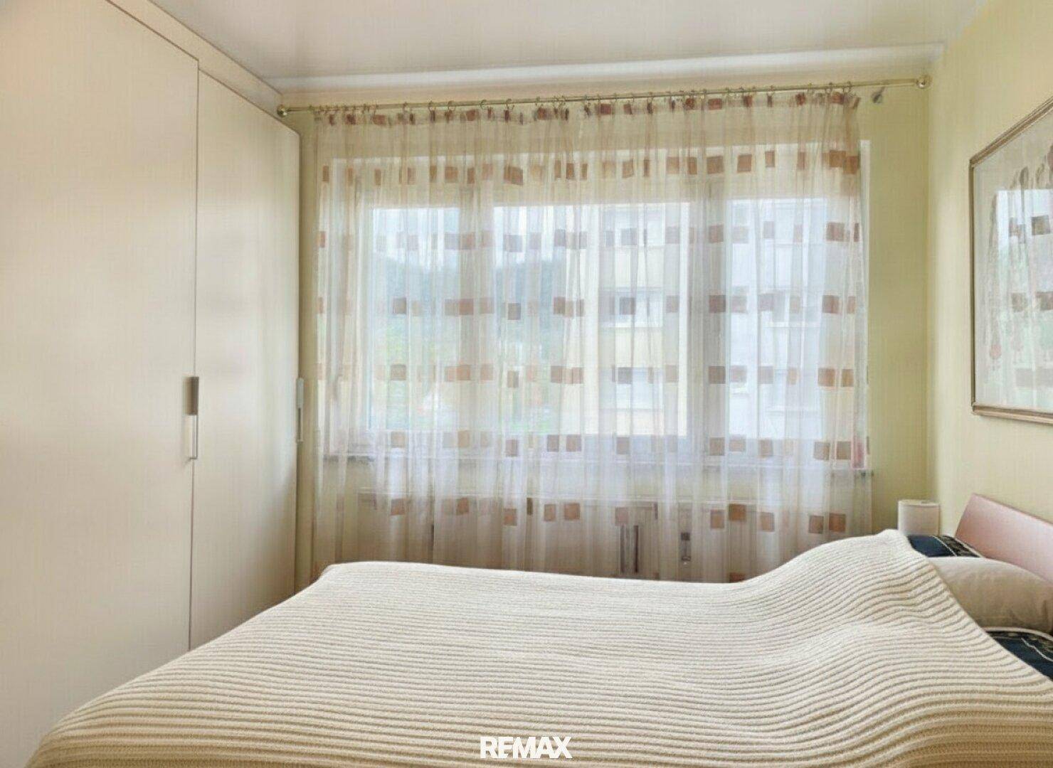 Helles Schlafzimmer mit Doppelbett, Kleiderschrank und Fenster mit Vorhängen.