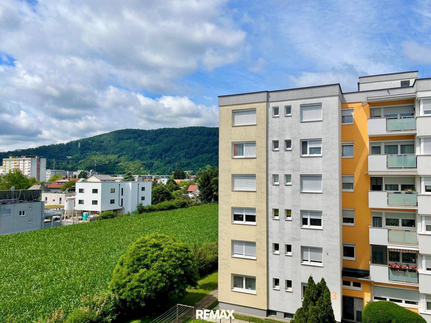 Mehrfamilienhausfassade mit Blick auf grüne Felder und bewaldete Hügel.