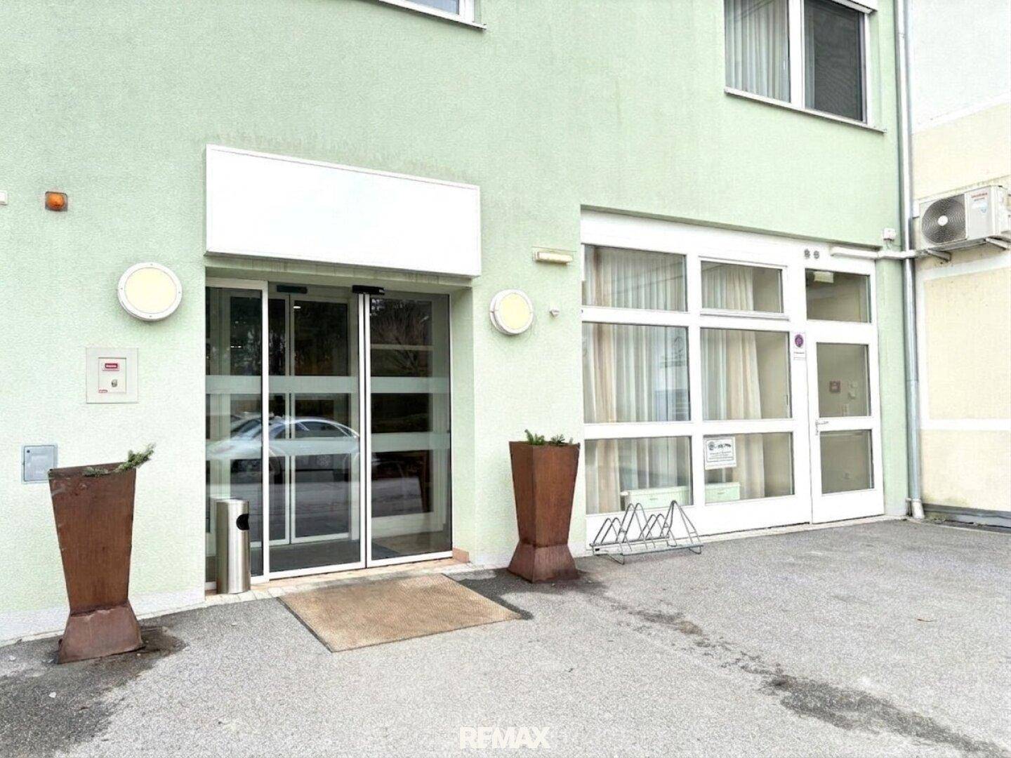 Eingangsbereich eines Gebäudes mit Glastüren, Fahrradständern und einer Klimaanlage an der Fassade.