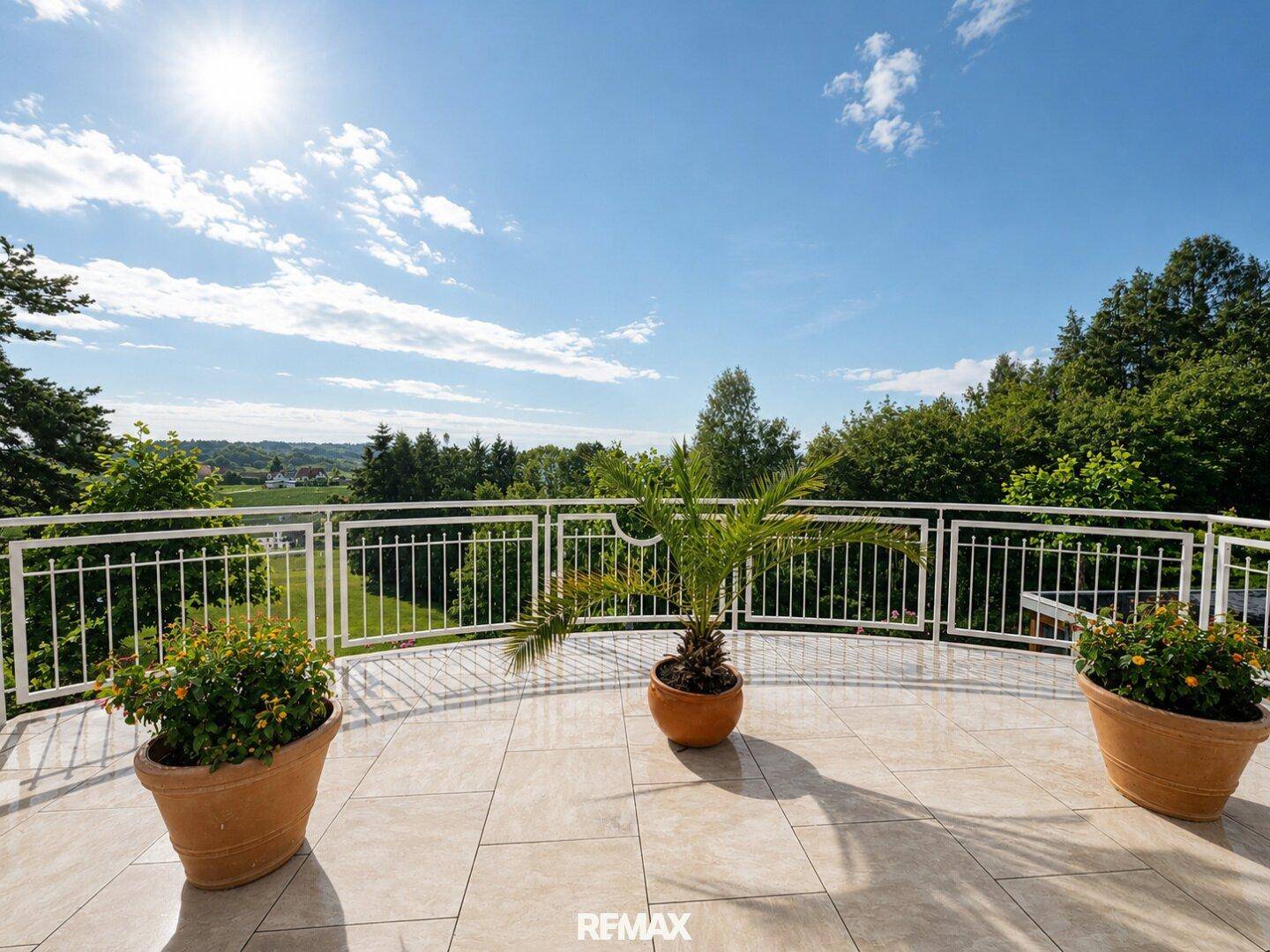 Großer Balkon mit Fliesenboden, Pflanzen und weitem Blick über die grüne Landschaft unter blauem Himmel.