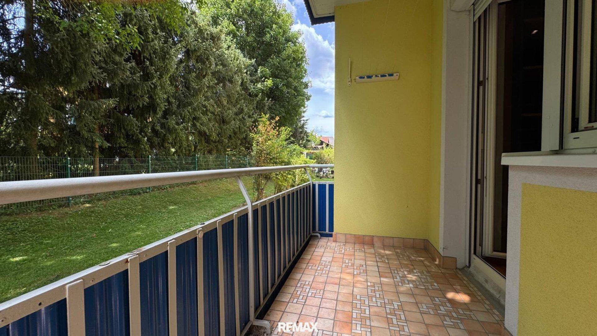 Balkon mit Fliesenboden und weitem Blick ins Grüne, umgeben von hohen Bäumen.