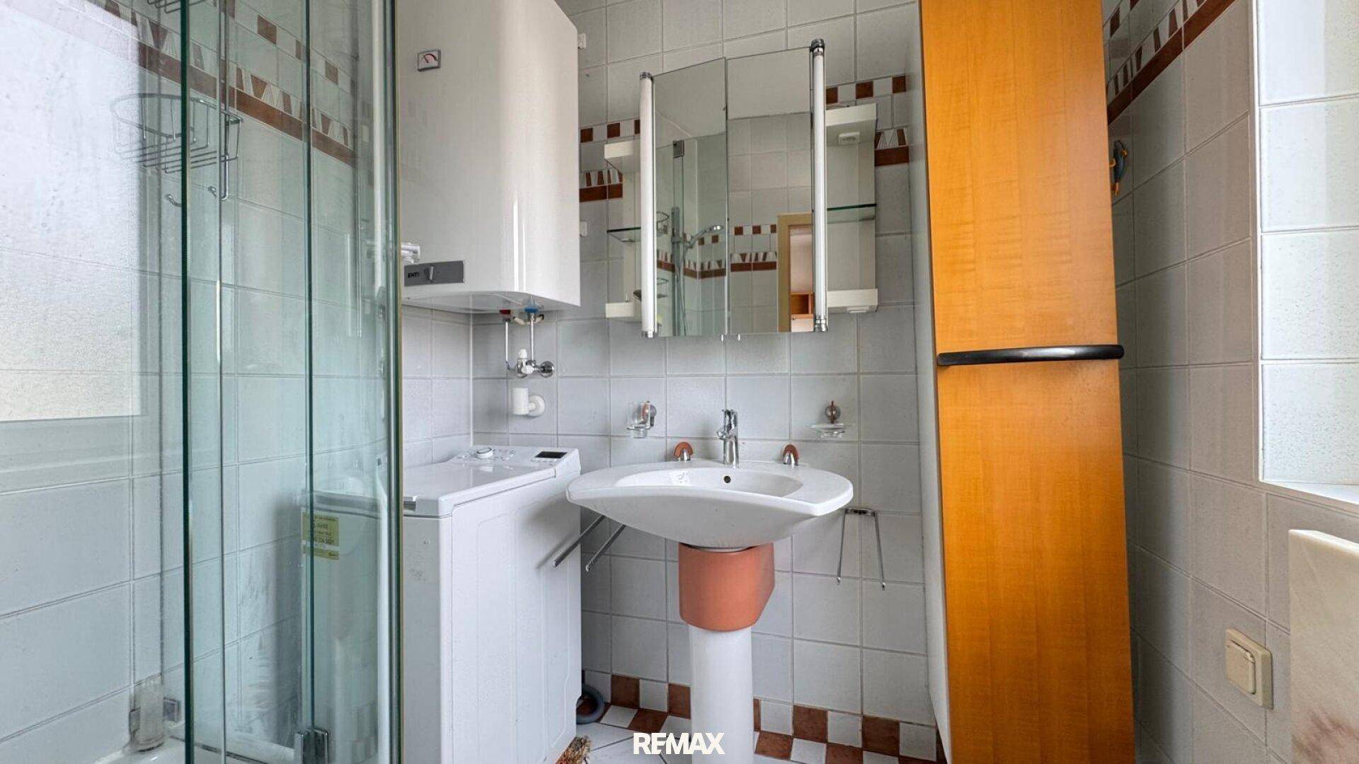 Badezimmer mit Dusche, Waschbecken, Spiegelschrank, Waschmaschine und 80er-Jahre-Fliesen.