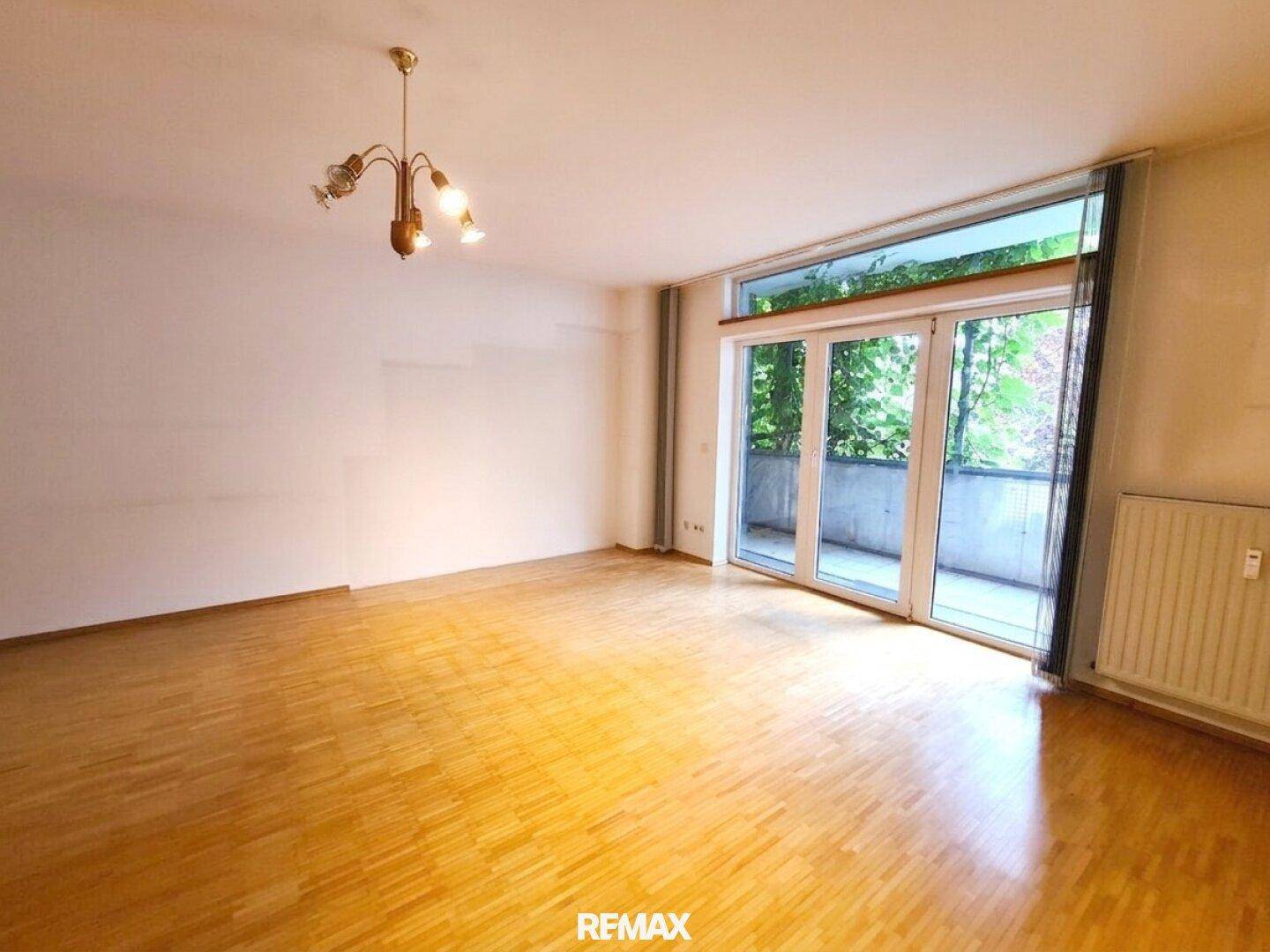 Leeres Wohnzimmer mit Parkettboden und großen Glastüren, die zu einem Balkon oder einer Terrasse führen.