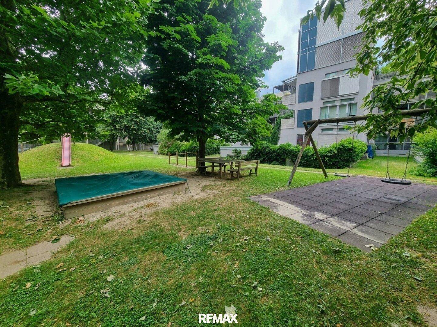 Gemeinschaftlicher Gartenbereich mit Spielplatz, Sandkasten, Schaukeln und Rutsche, umgeben von Bäumen.