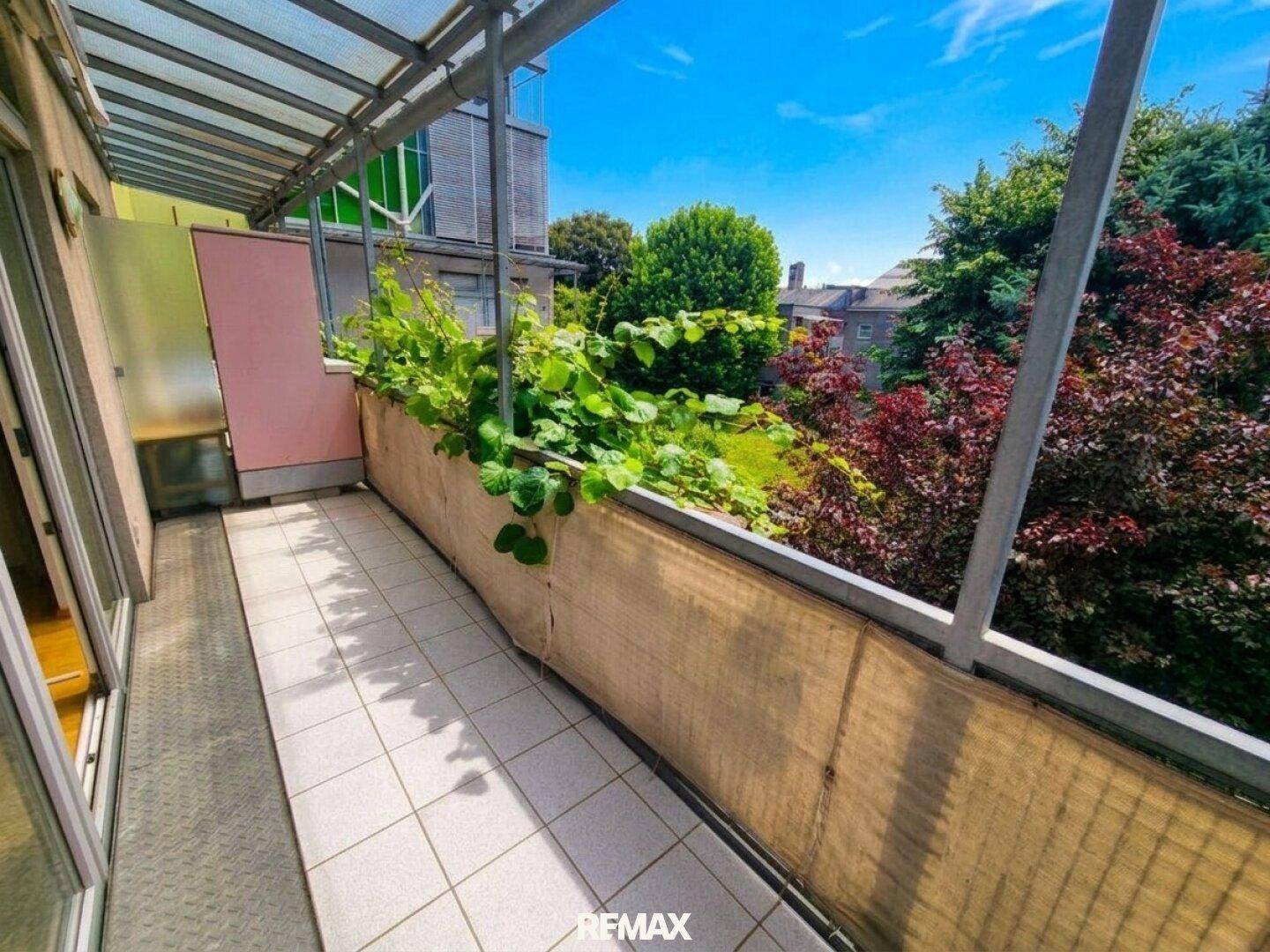 Heller Balkon mit Fliesenboden, Pflanzenkübeln und Blick ins Grüne, überdacht mit einem Glasdach.