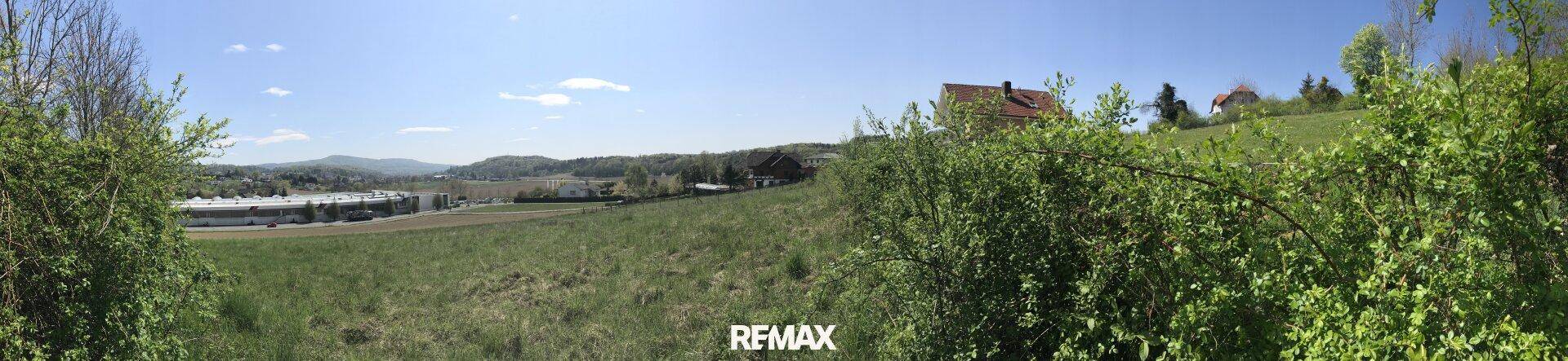 Weitläufiger Panoramablick über grüne Felder und Büsche mit Gebäuden in der Ferne.