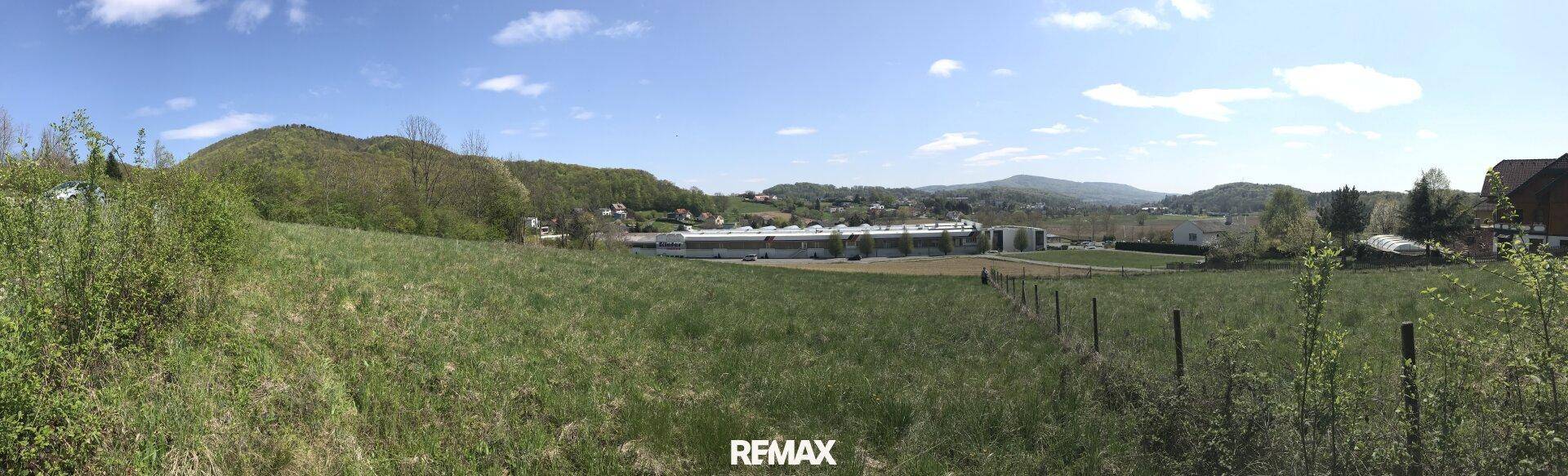 Breiter Panoramablick über grüne Wiesen mit bewaldeten Hügeln und Gebäuden am Horizont.