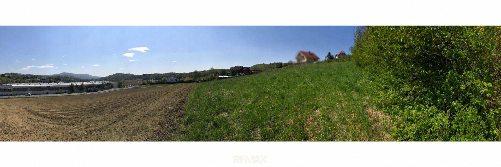 Panoramablick auf eine Landschaft mit grünen Wiesen, Ackerland und entfernten Gebäuden unter blauem Himmel.
