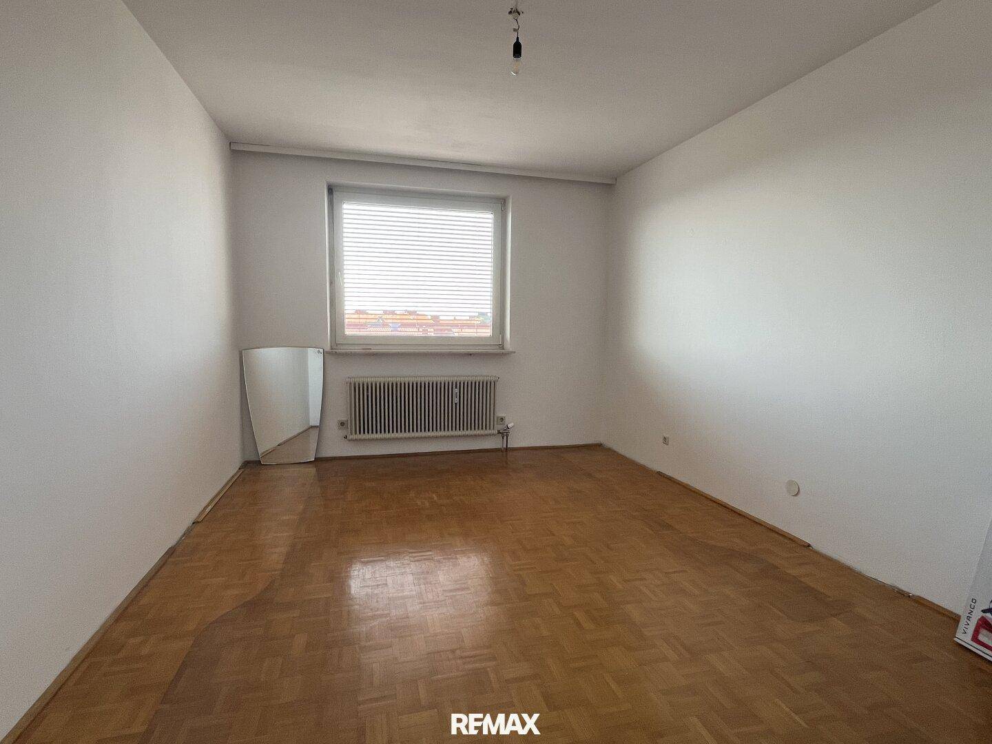 Leeres Schlafzimmer mit Parkettboden, einem Fenster und einem Heizkörper.