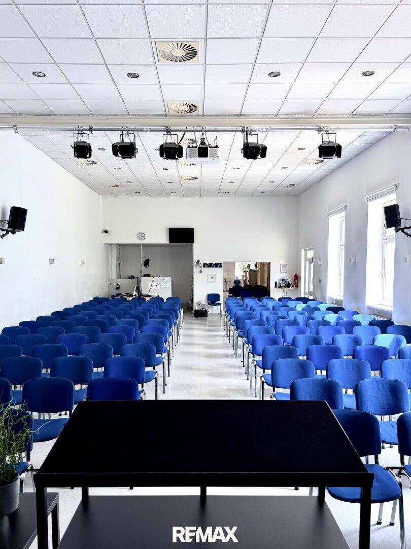 Blick vom Podium in den Konferenzraum mit blauen Stühlen, hohen Decken und Deckenlüftung.