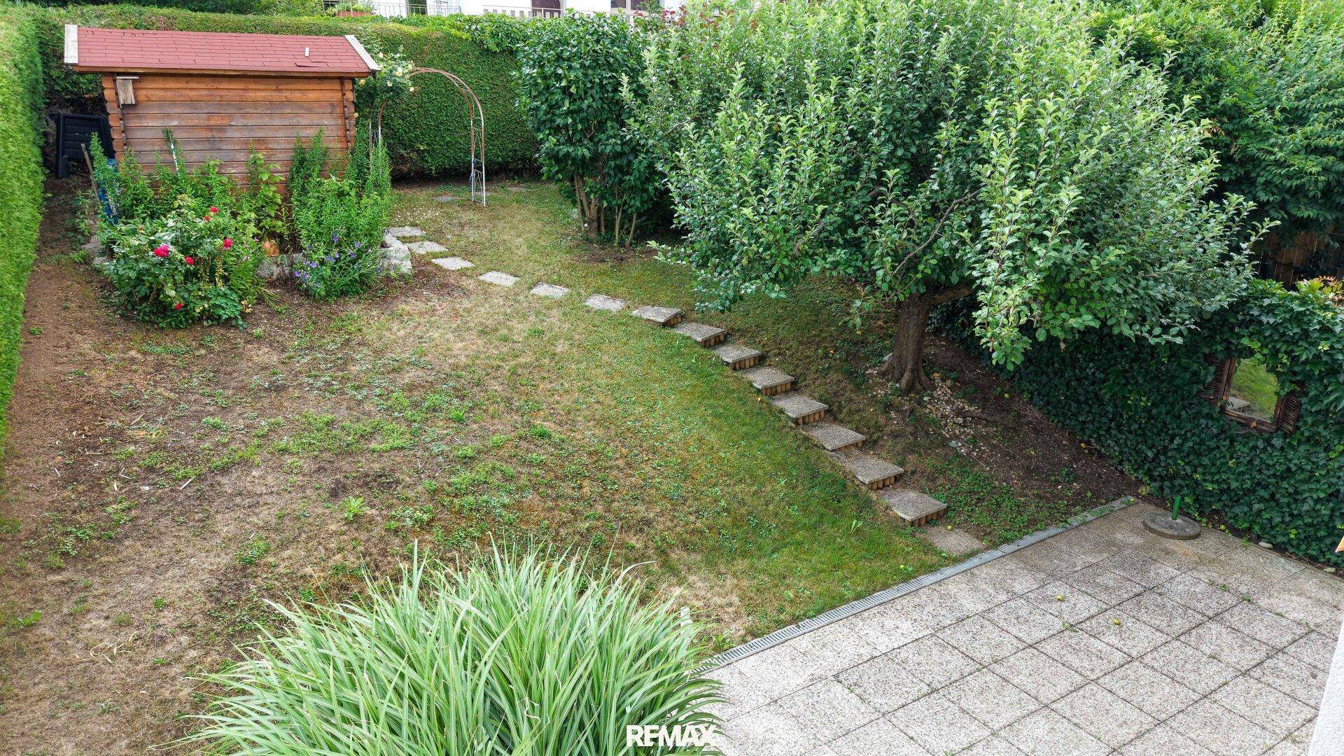 Weitläufiger Gartenbereich mit Rasenfläche, Gartenhaus und Trittsteinen, umgeben von Hecken.