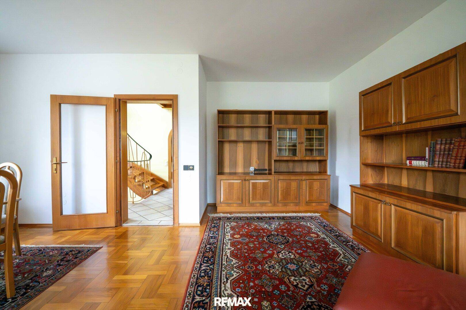 Wohnbereich mit Parkettboden, Holzschränken und Zugang zum Flur mit Treppe.