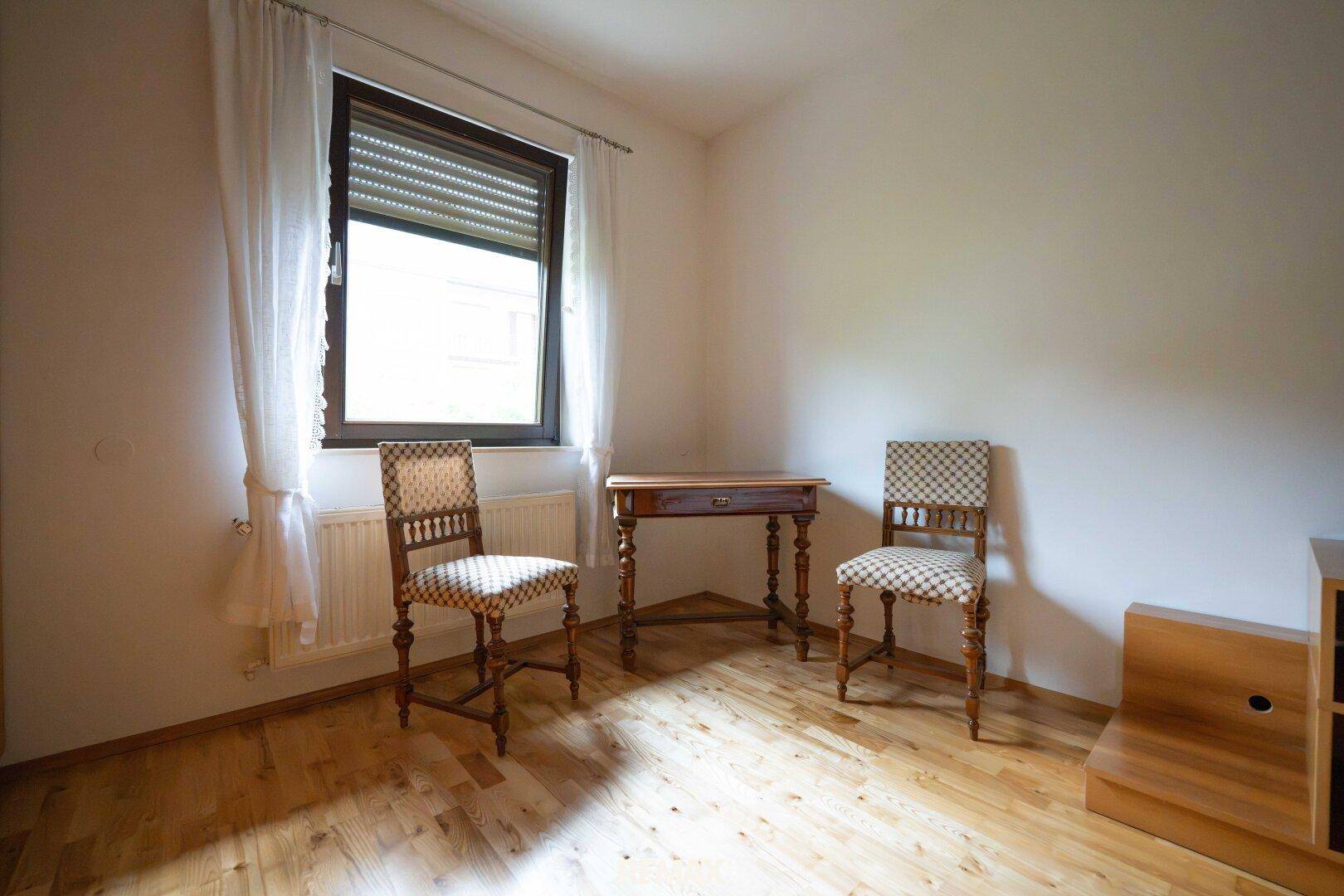 Helles Zimmer mit Holzboden, zwei Stühlen und einem Fenster mit Rollladen.