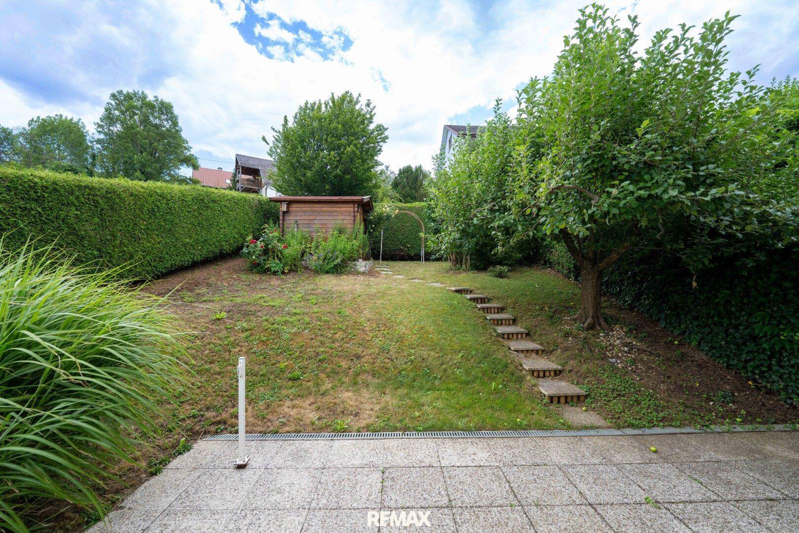 Gepflegter Gartenbereich mit Rasenfläche, Hecke, Gartenhaus und Steinstufen.