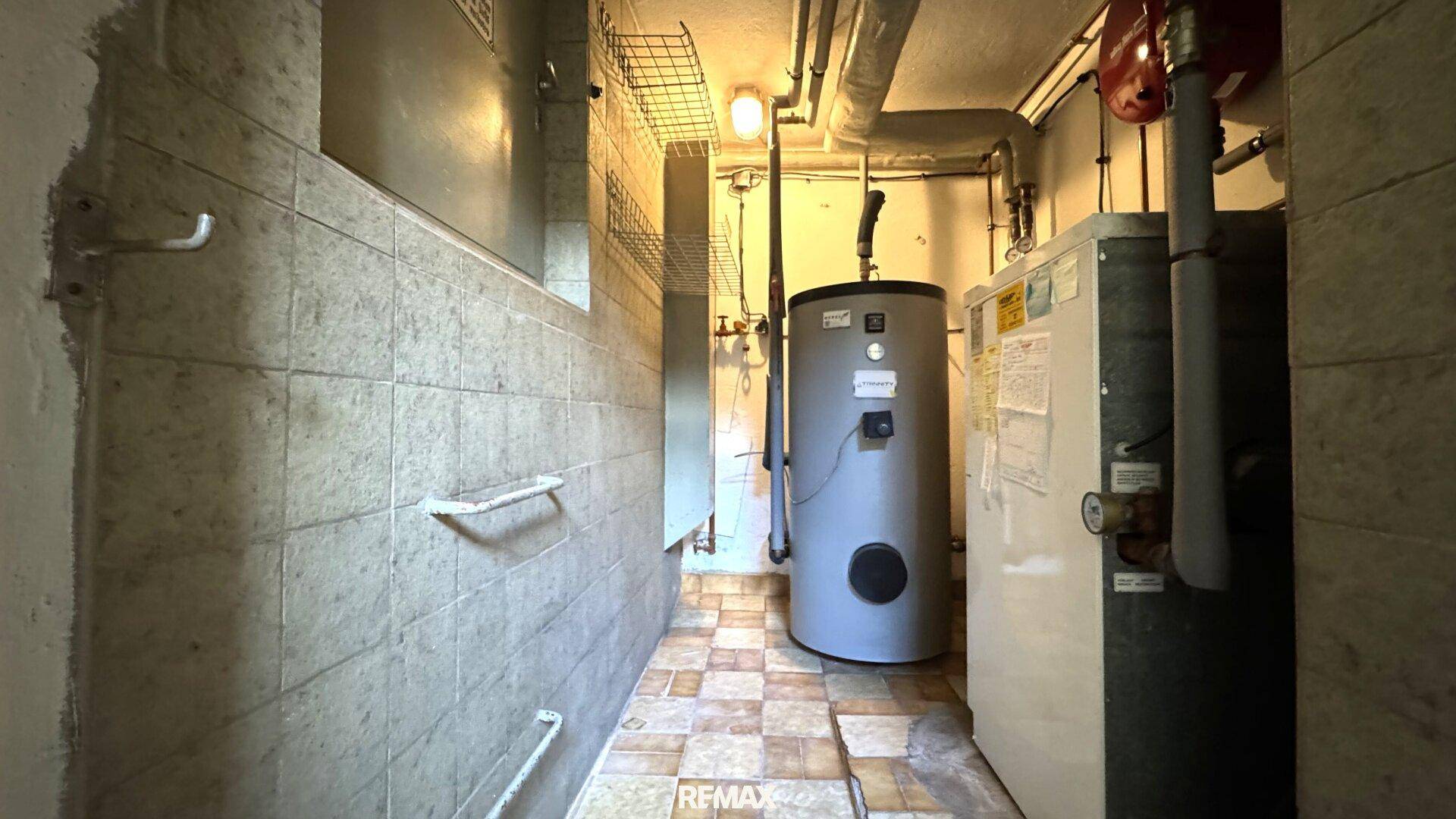 Funktionaler Technikraum mit Warmwasserboiler, sichtbaren Leitungen und robusten Fliesenwänden.
