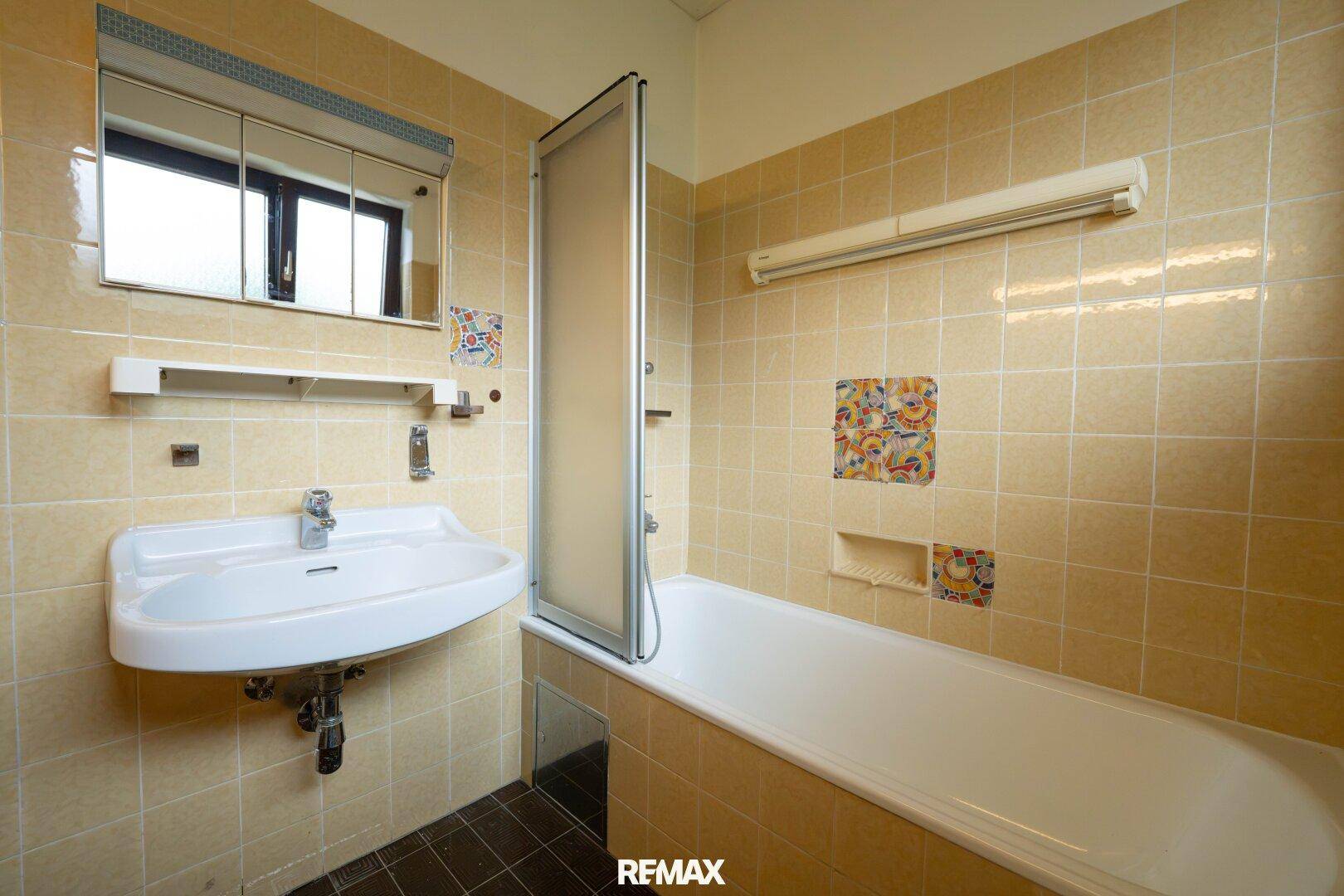 Badezimmer mit beigefarbenen Fliesen, Waschbecken, Badewanne und Spiegelschrank.