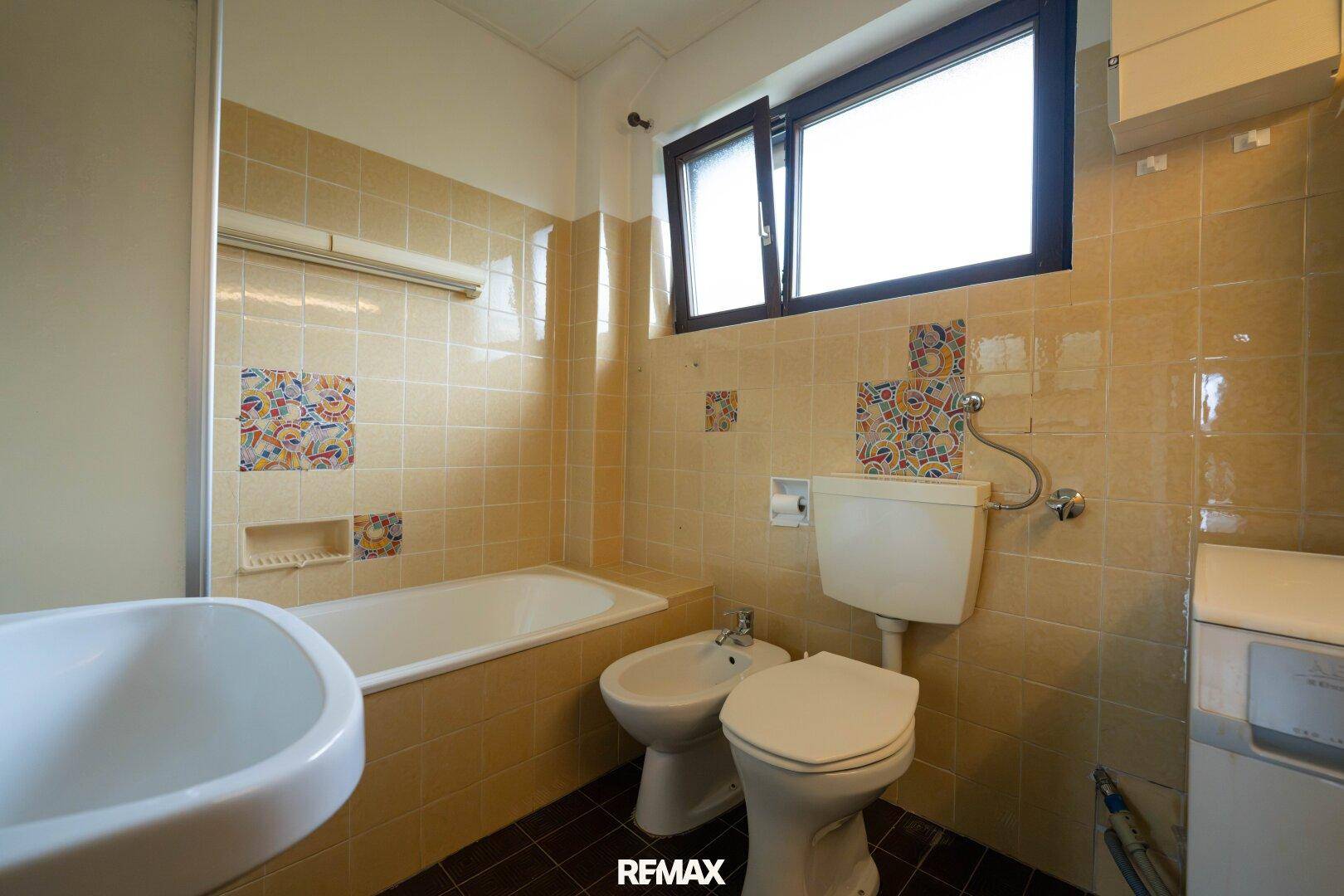 Badezimmer mit beigefarbenen Fliesen, Badewanne, Bidet und Fenster für Tageslicht.