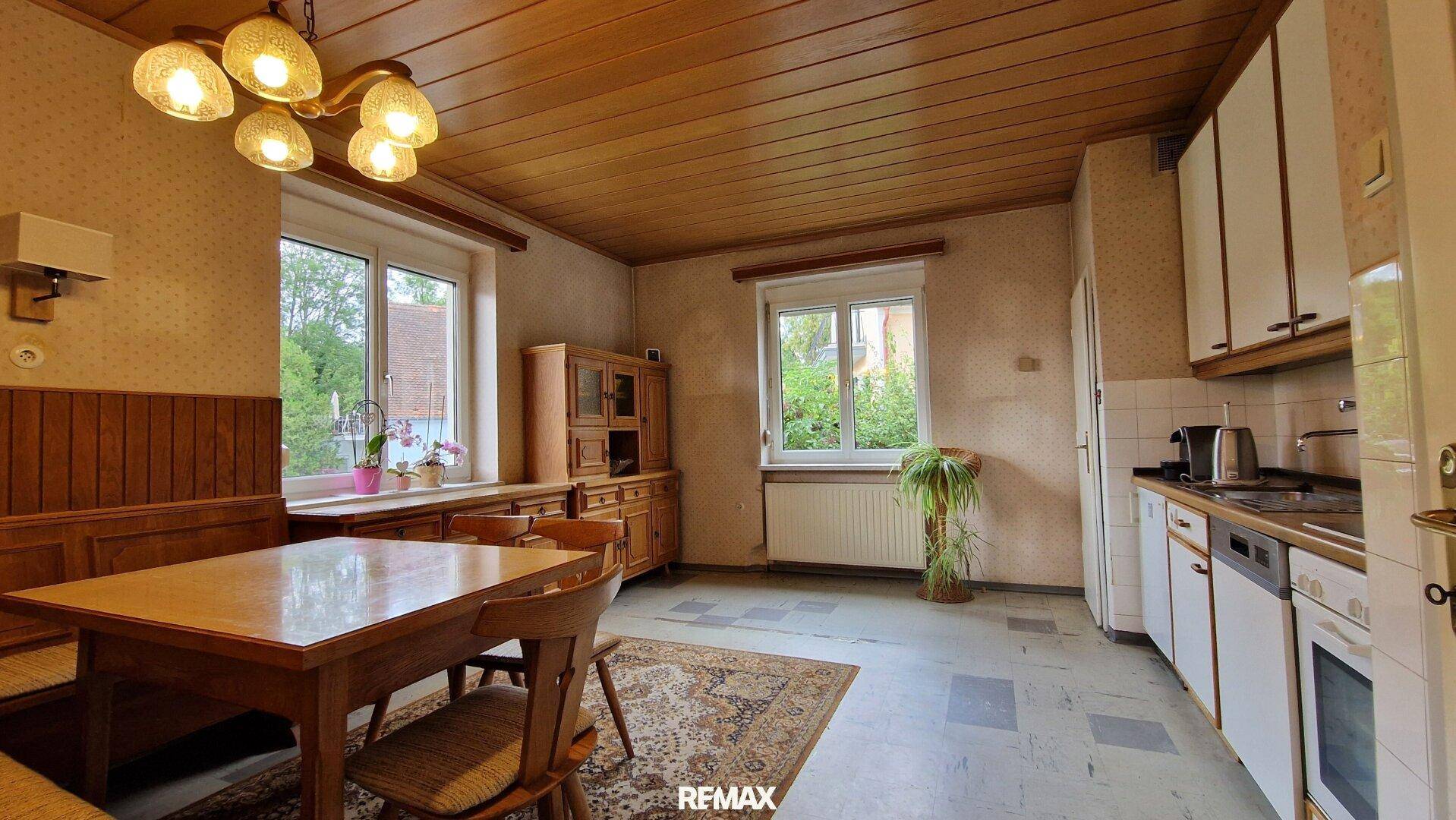 Geräumige Wohnküche mit Holzdecke, traditionellen Möbeln und Blick ins Grüne durch die Fenster.