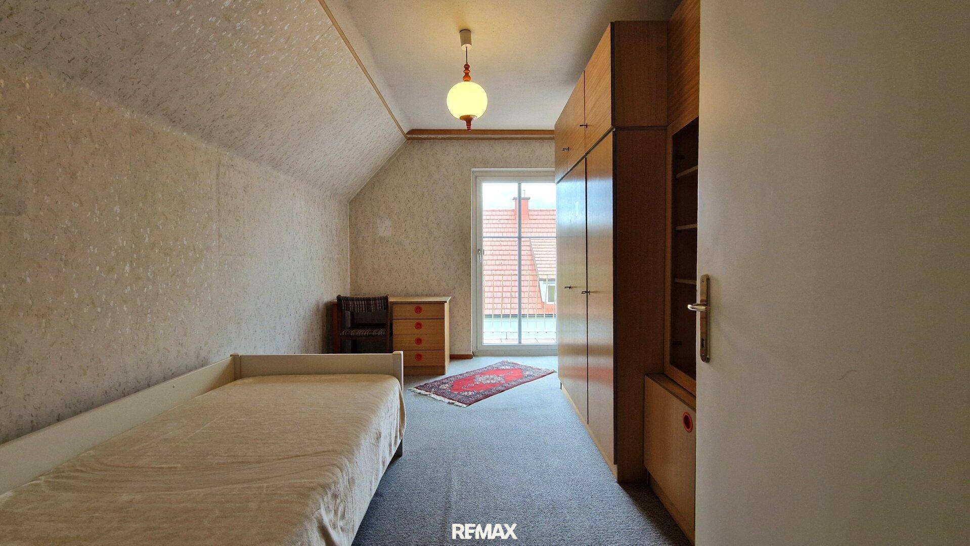 Helles Dachgeschoss-Schlafzimmer mit Einzelbett, Schrankwand und Blick nach draußen durch das Fenster.