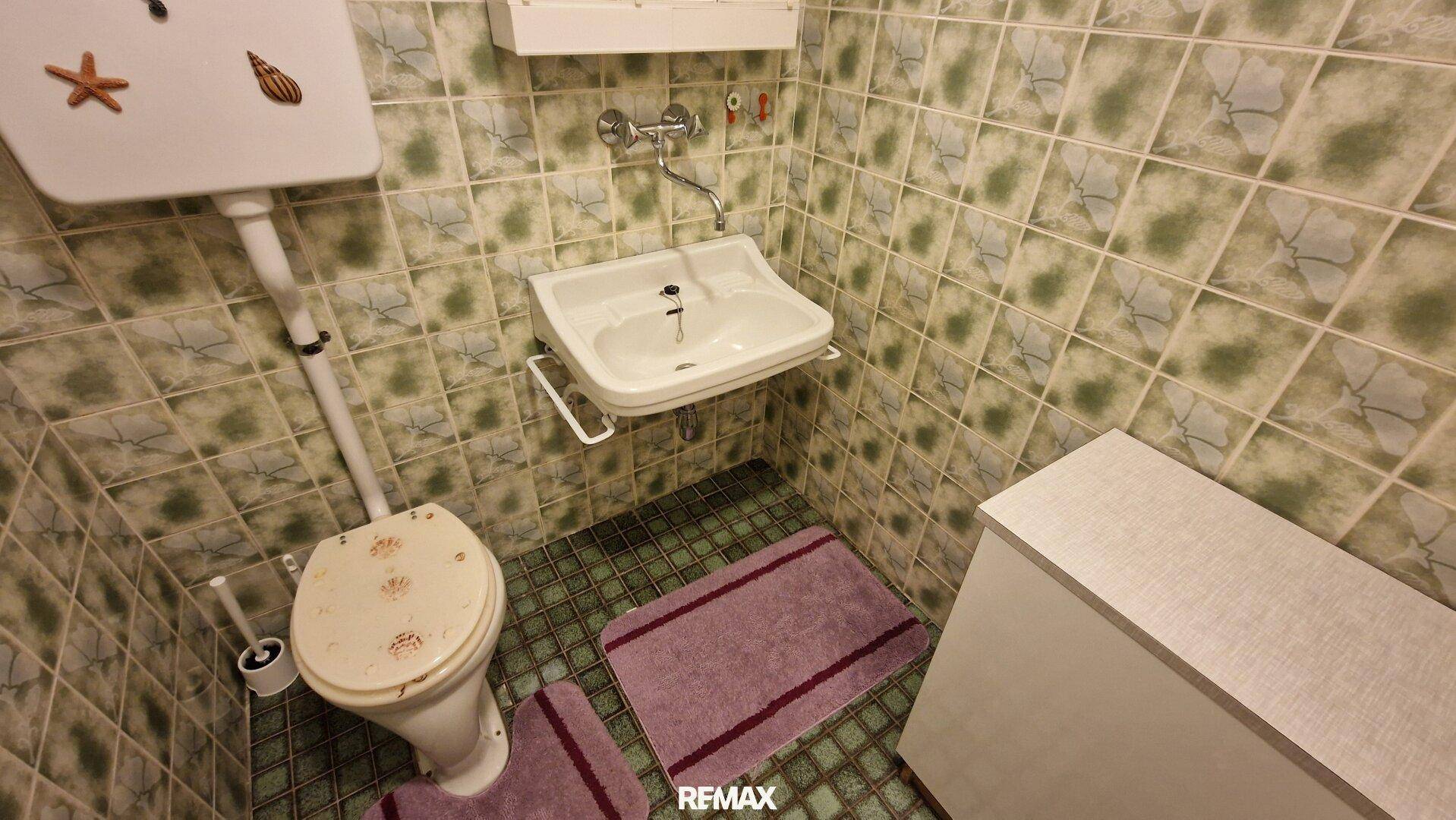Einfaches WC mit grün gemusterten Fliesen, kleinem Waschbecken und einem Spülkasten.