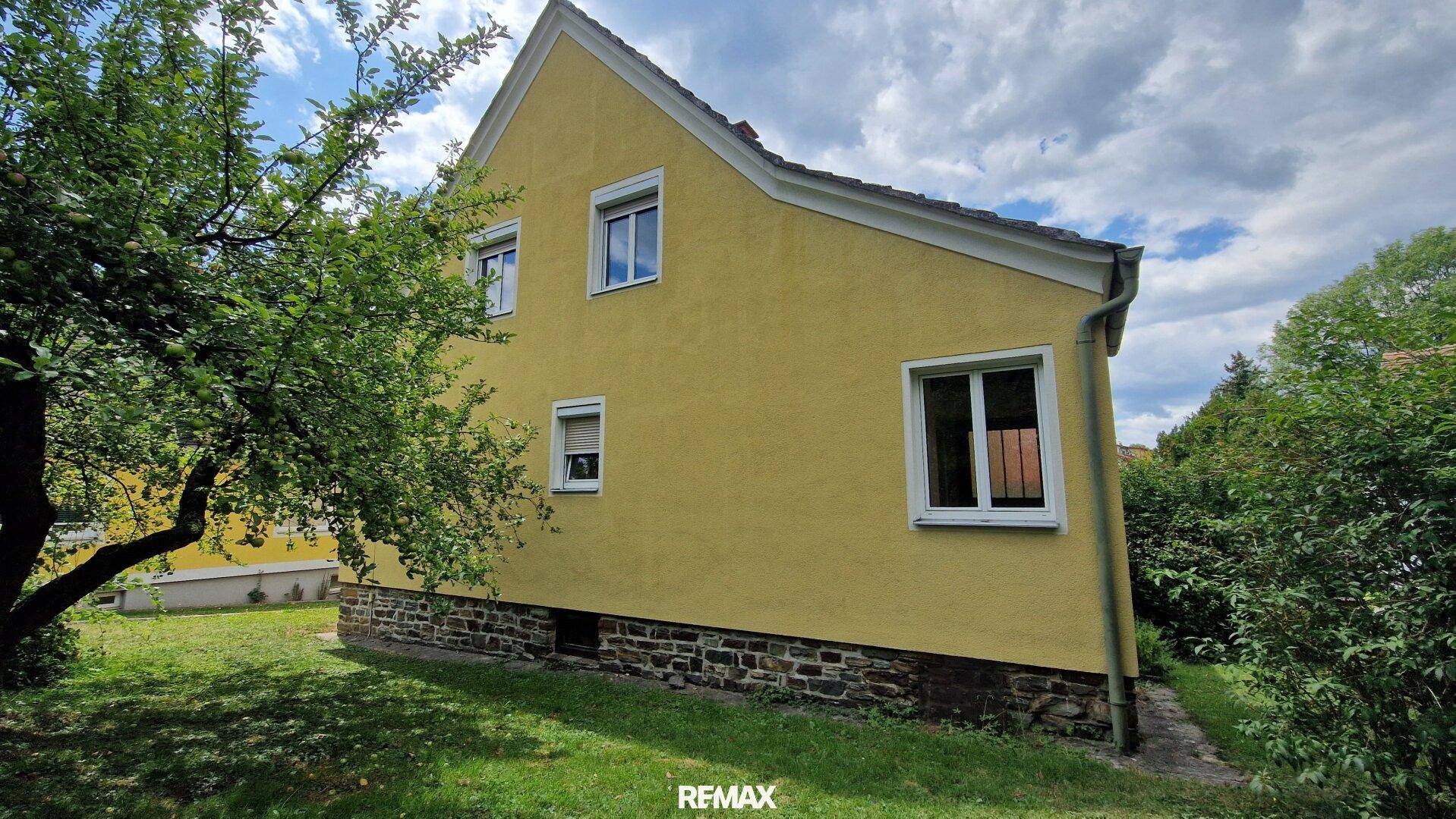 Seitenansicht des Hauses mit gelber Fassade, weißen Fenstern und einem robusten Steinsockel im Grünen.