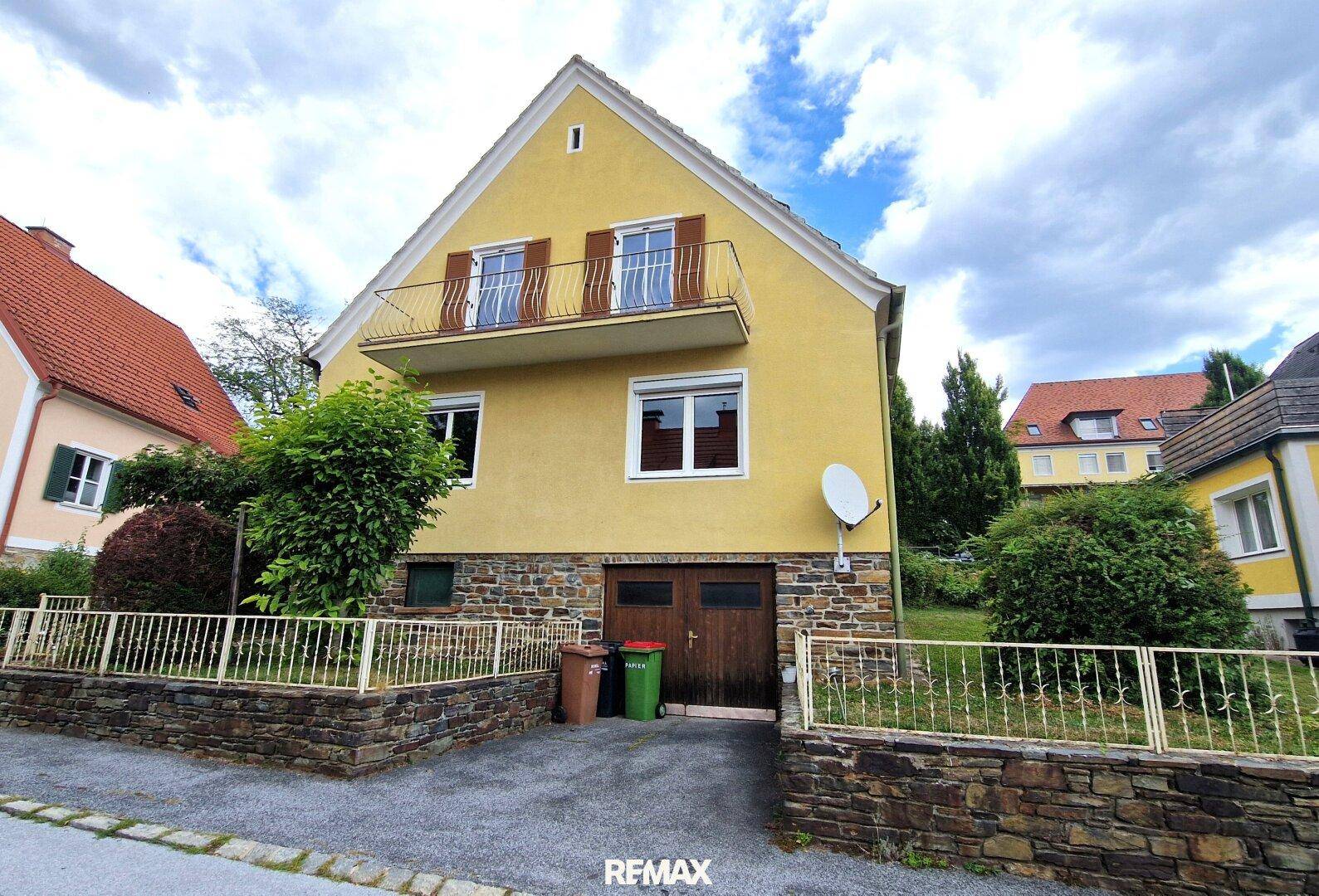 Vorderansicht des gelben Hauses mit Balkon, integrierter Garage und einer gepflasterten Auffahrt.