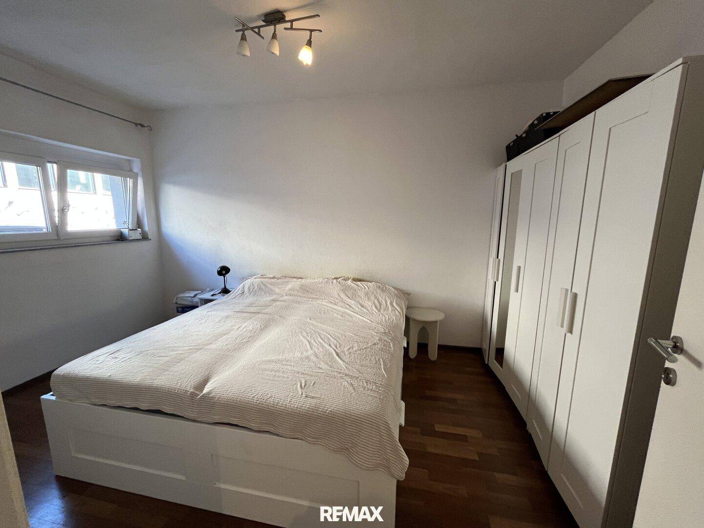 Schlafzimmer mit Doppelbett, weißem Kleiderschrank und Holzparkettboden, gut beleuchtet durch ein Fenster.