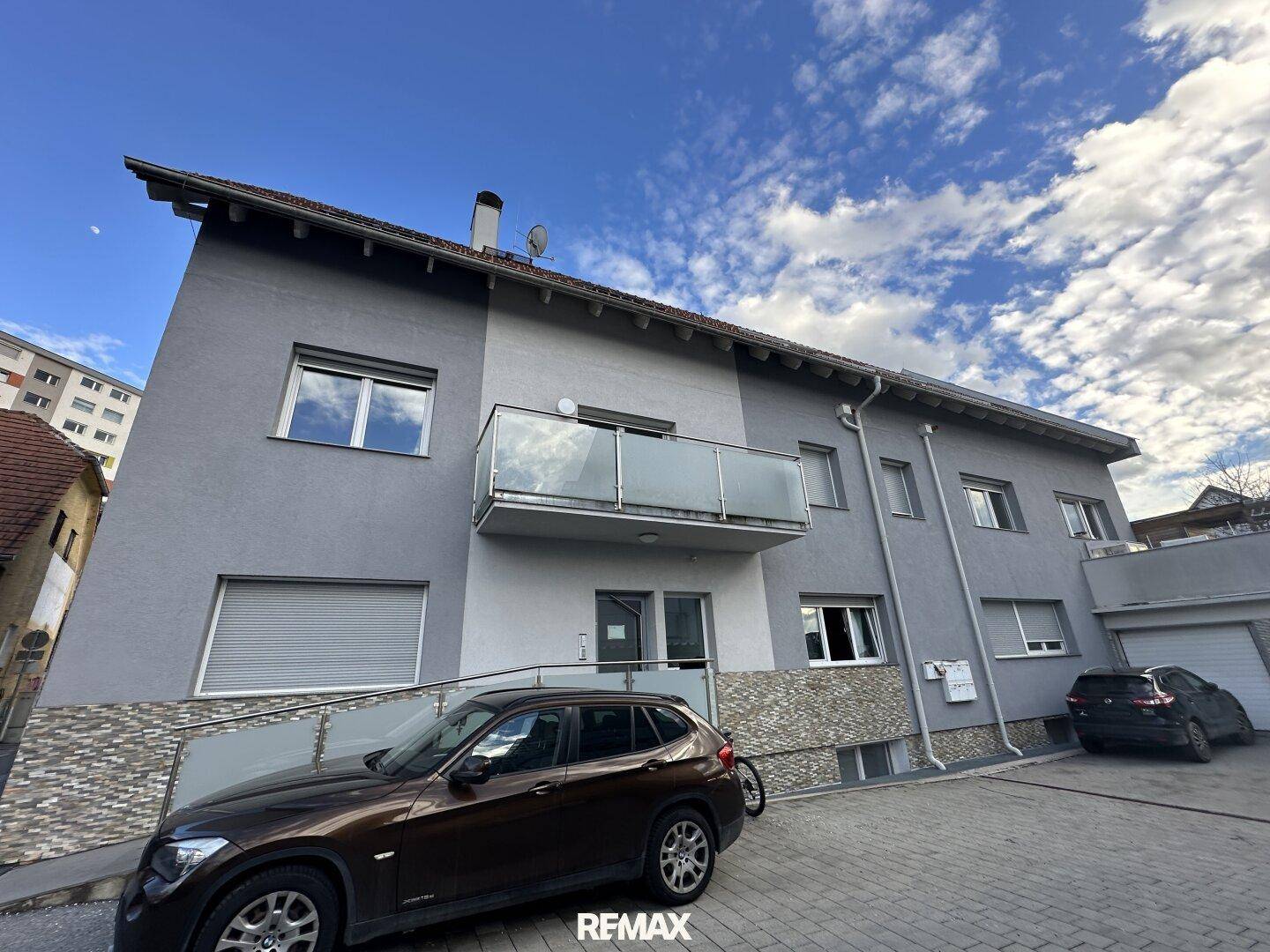 Modernes Mehrfamilienhaus mit grauer Fassade, Balkon und Rollläden an den Fenstern.