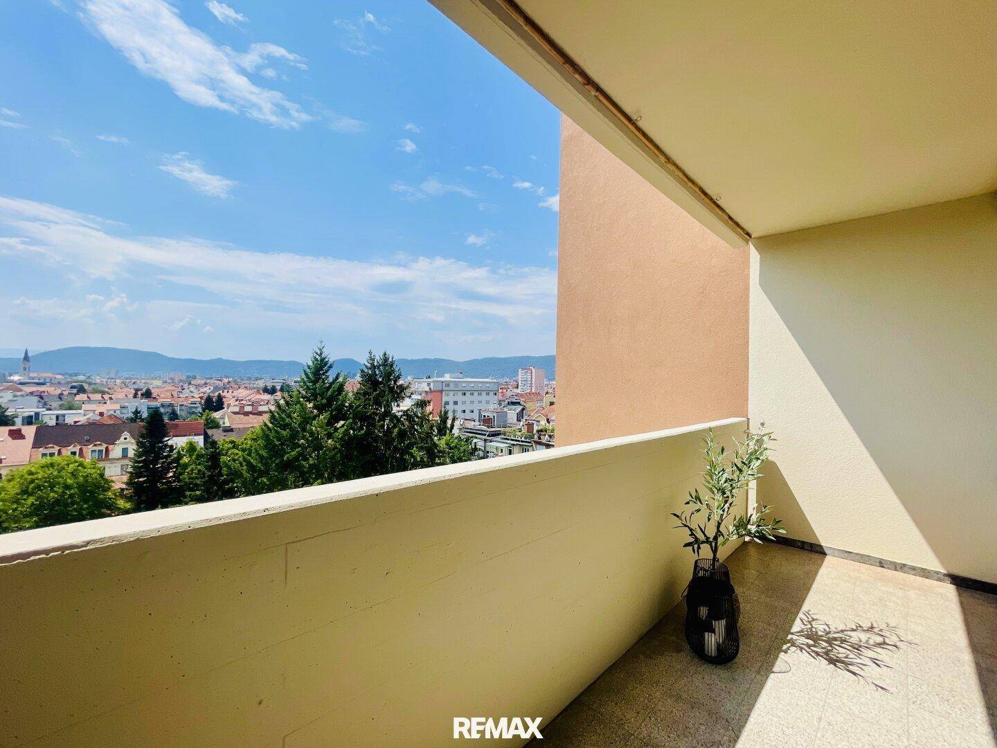 Sonniger Balkon mit Stadtblick und grüner Umgebung unter blauem Himmel.