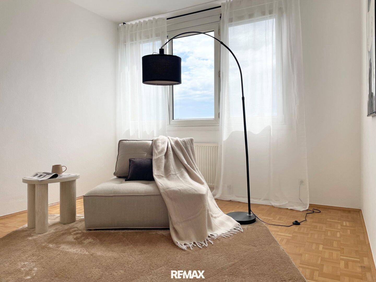 Heller Wohnbereich mit beigem Sessel, Stehlampe und Fenster mit Vorhängen.