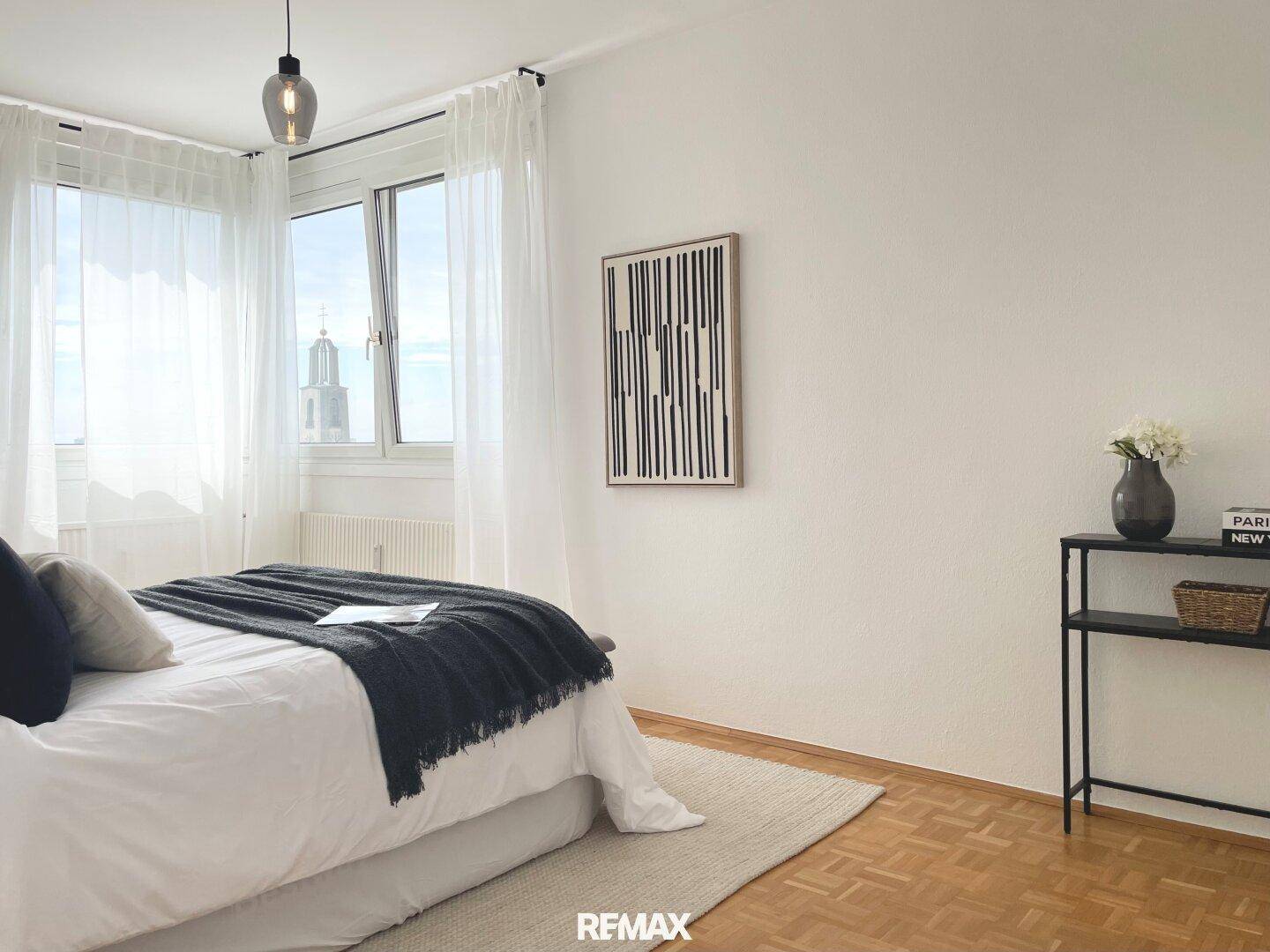 Helles Schlafzimmer mit Doppelbett, Parkettboden und Fenster mit Stadtblick.