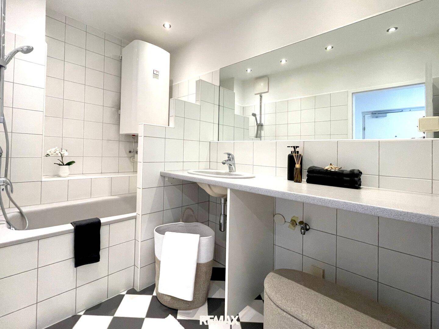 Helles Badezimmer mit Badewanne, Duschfunktion, Waschbecken und einem markanten schwarz-weiß karierten Fliesenboden.