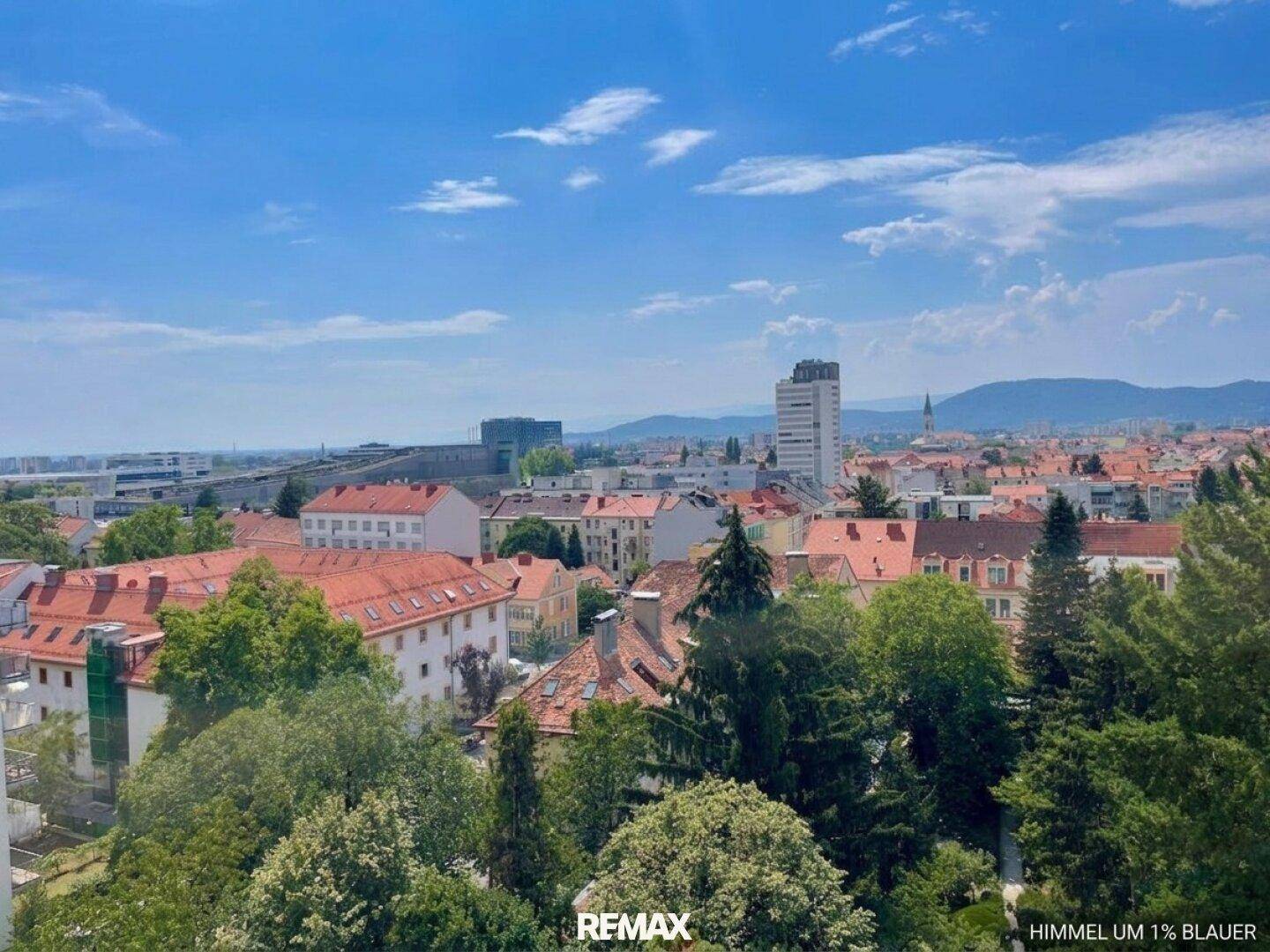 Weitläufiger Stadtblick mit grünen Bäumen, Wohngebäuden und einem klaren blauen Himmel.