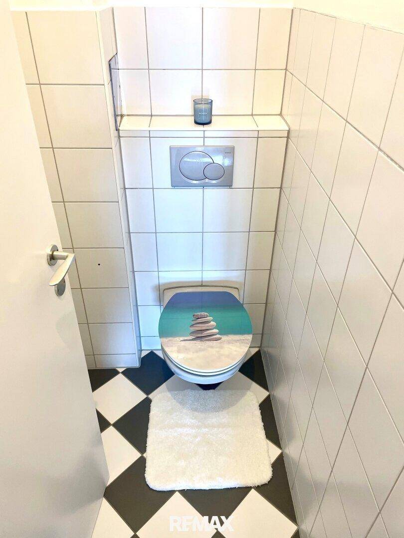 Separates WC mit weiß gefliesten Wänden, einem dekorativen Toilettensitz und schwarz-weiß kariertem Boden.