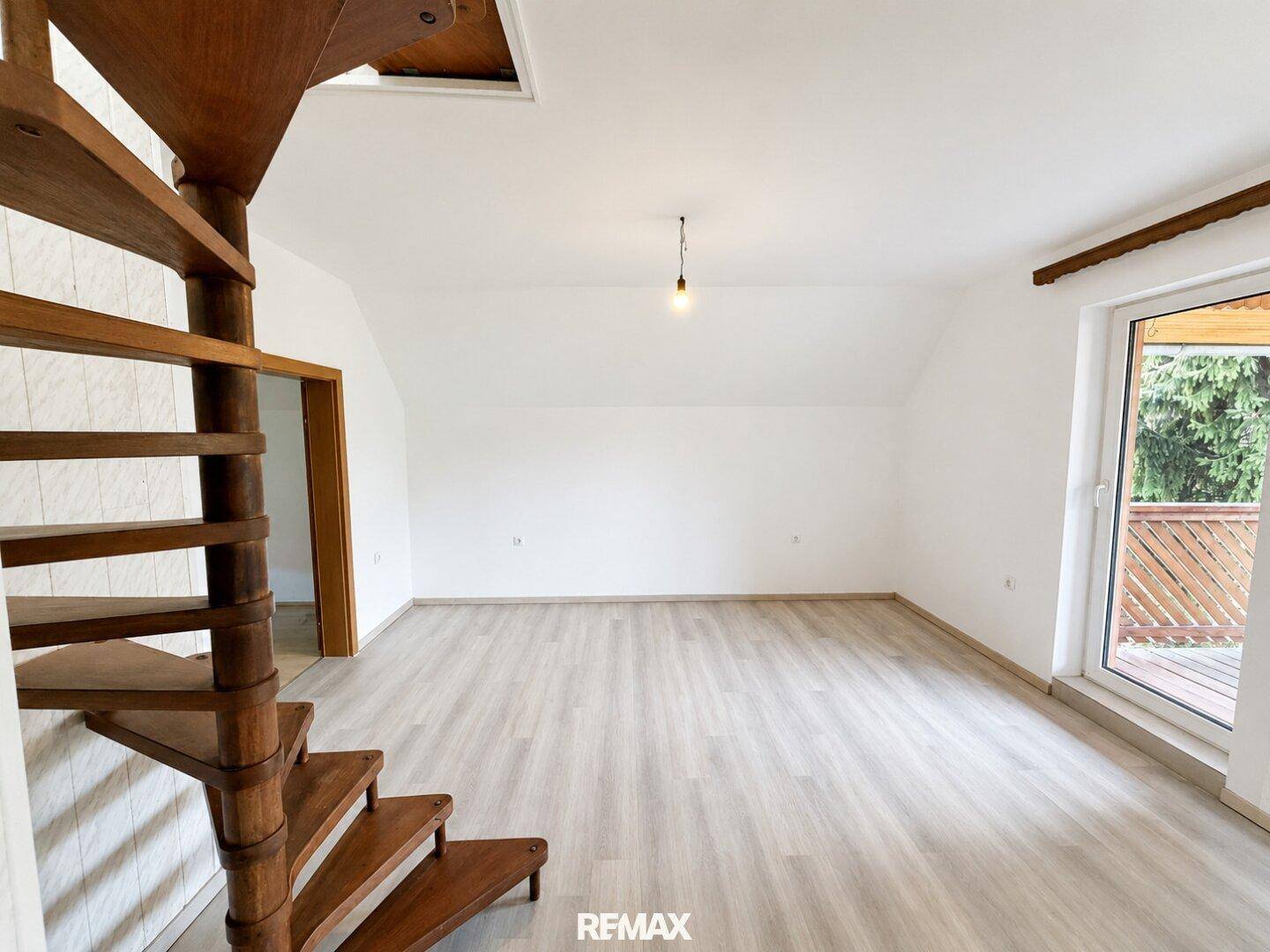 Geräumiges, leeres Wohnzimmer mit Laminatboden, Holztreppe und Balkonzugang.