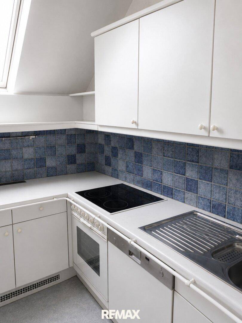 Küchenzeile mit weißen Schränken, integriertem Kochfeld, Backofen und Spülbecken unter einem Dachfenster.