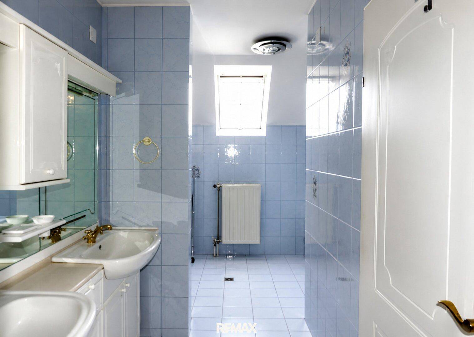 Badezimmer mit hellblauen Fliesen, Waschbecken, Spiegel und Dachfenster für Tageslicht.
