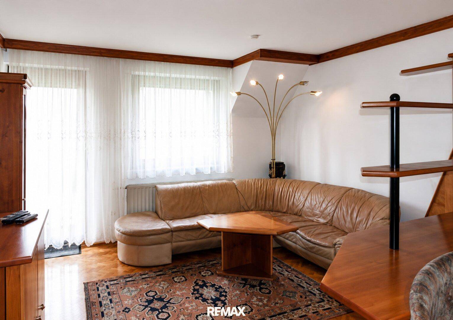 Heller Wohnbereich mit großem Ledersofa, Teppich und Fenstern mit Vorhängen.