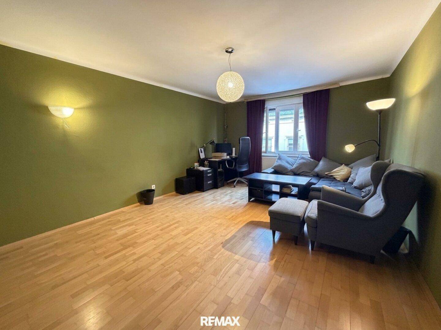 Gemütliches Wohnzimmer mit olivgrünen Wänden, hellem Parkettboden und einem Arbeitsbereich am Fenster.