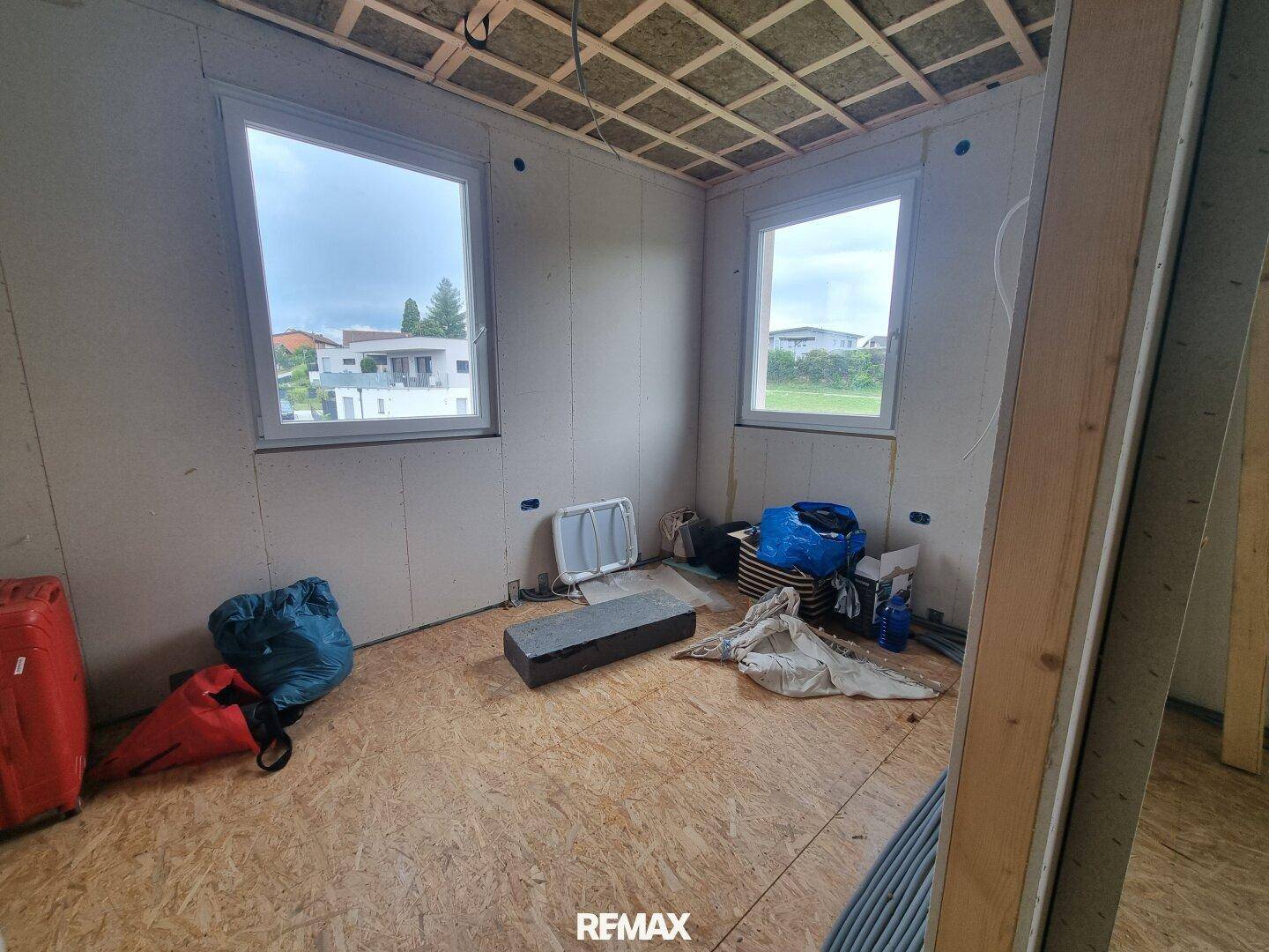 Rohbauzimmer mit zwei Fenstern und Holzspanplattenboden, mit Baumaterialien im Raum.