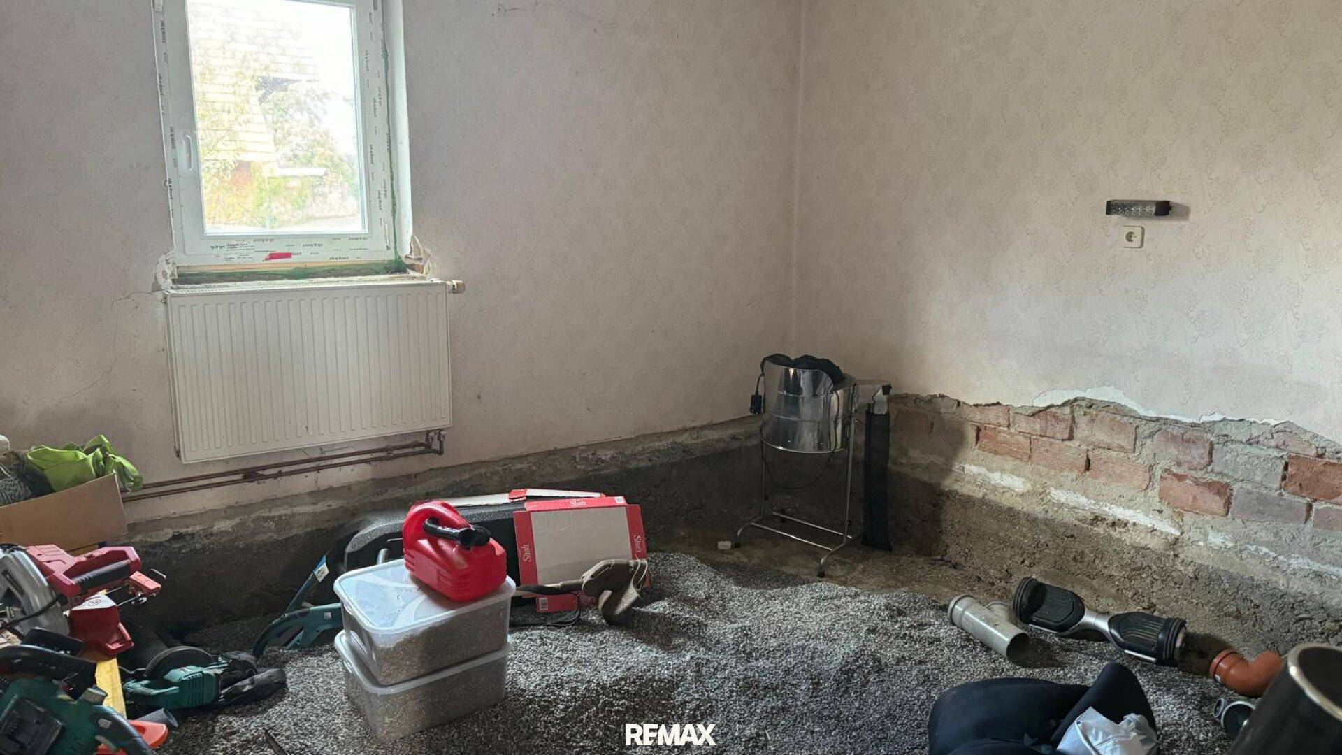 Raum in Renovierung mit freigelegten Wänden, Kiesboden und einem Fenster mit Heizkörper, bereit für den Ausbau.
